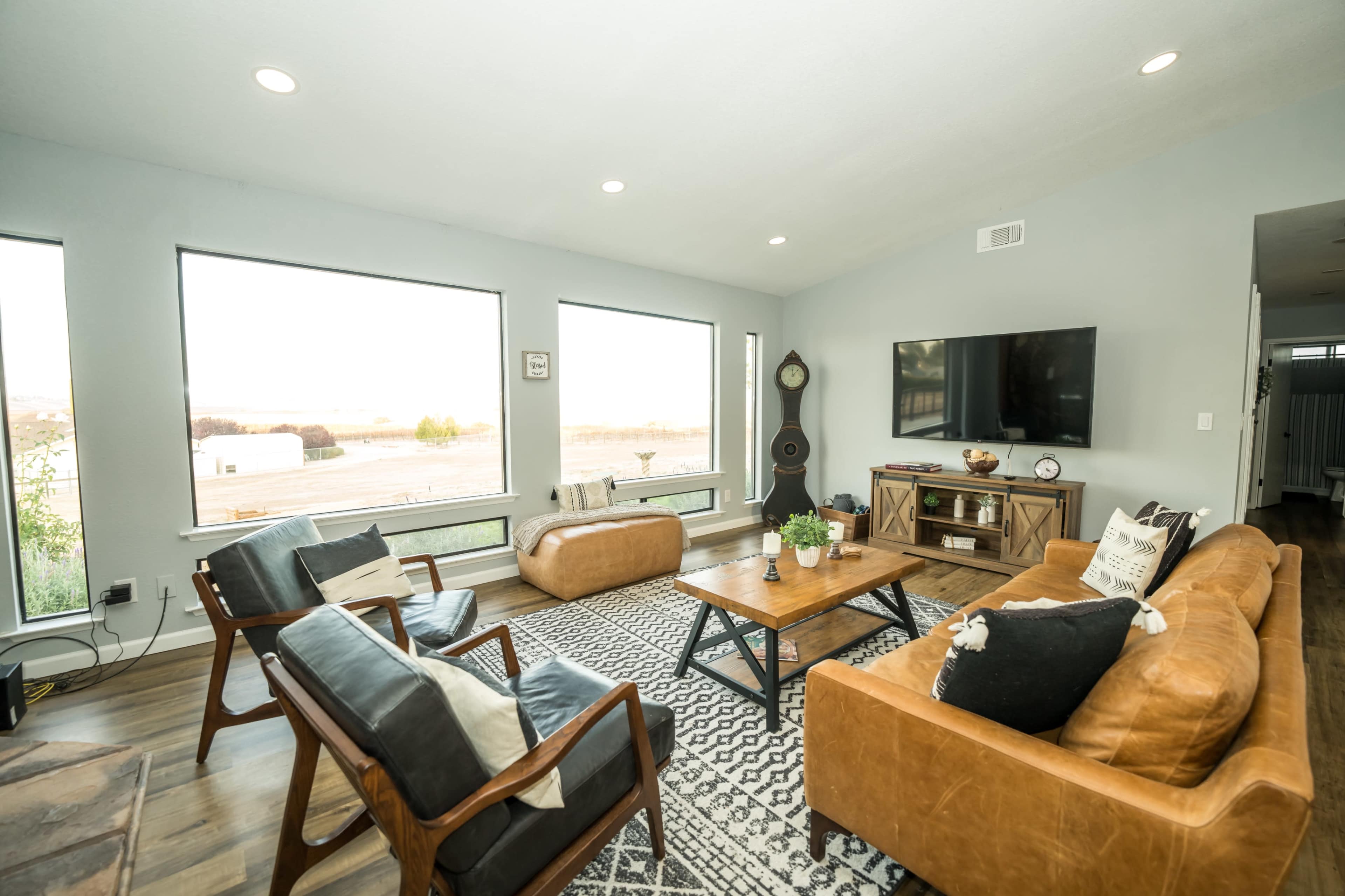 A modern living room features large windows with a view, leather and wooden furniture, and a flat-screen TV mounted on the wall.
