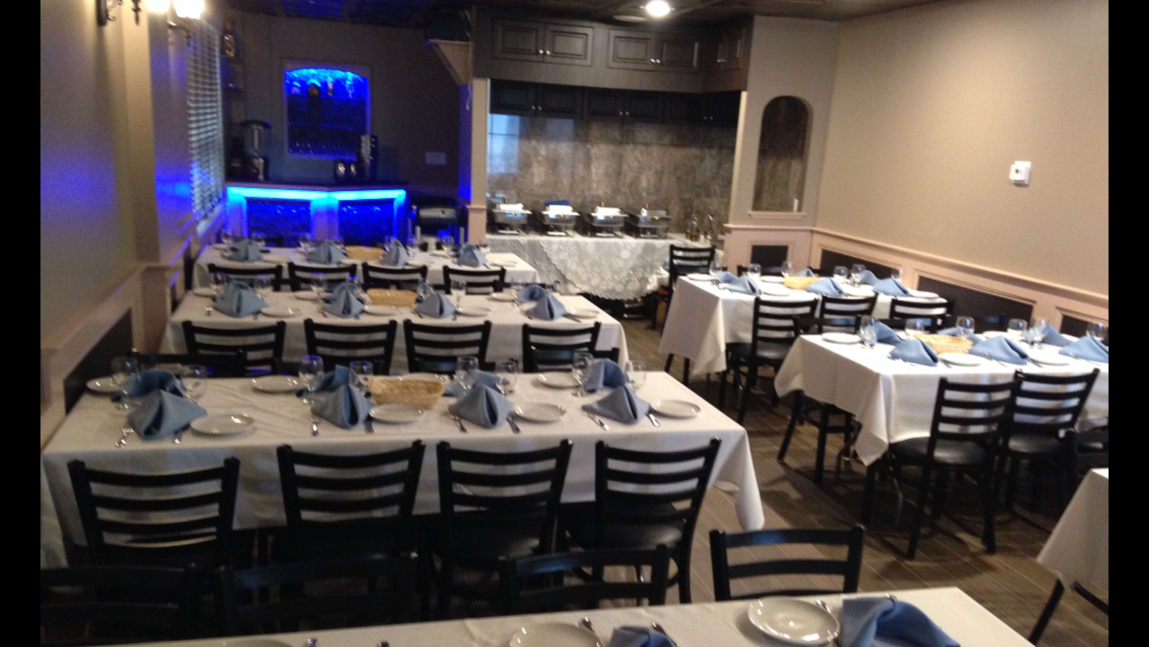 A dining room set with multiple tables dressed in white tablecloths and blue napkins, with a bar area in the background.