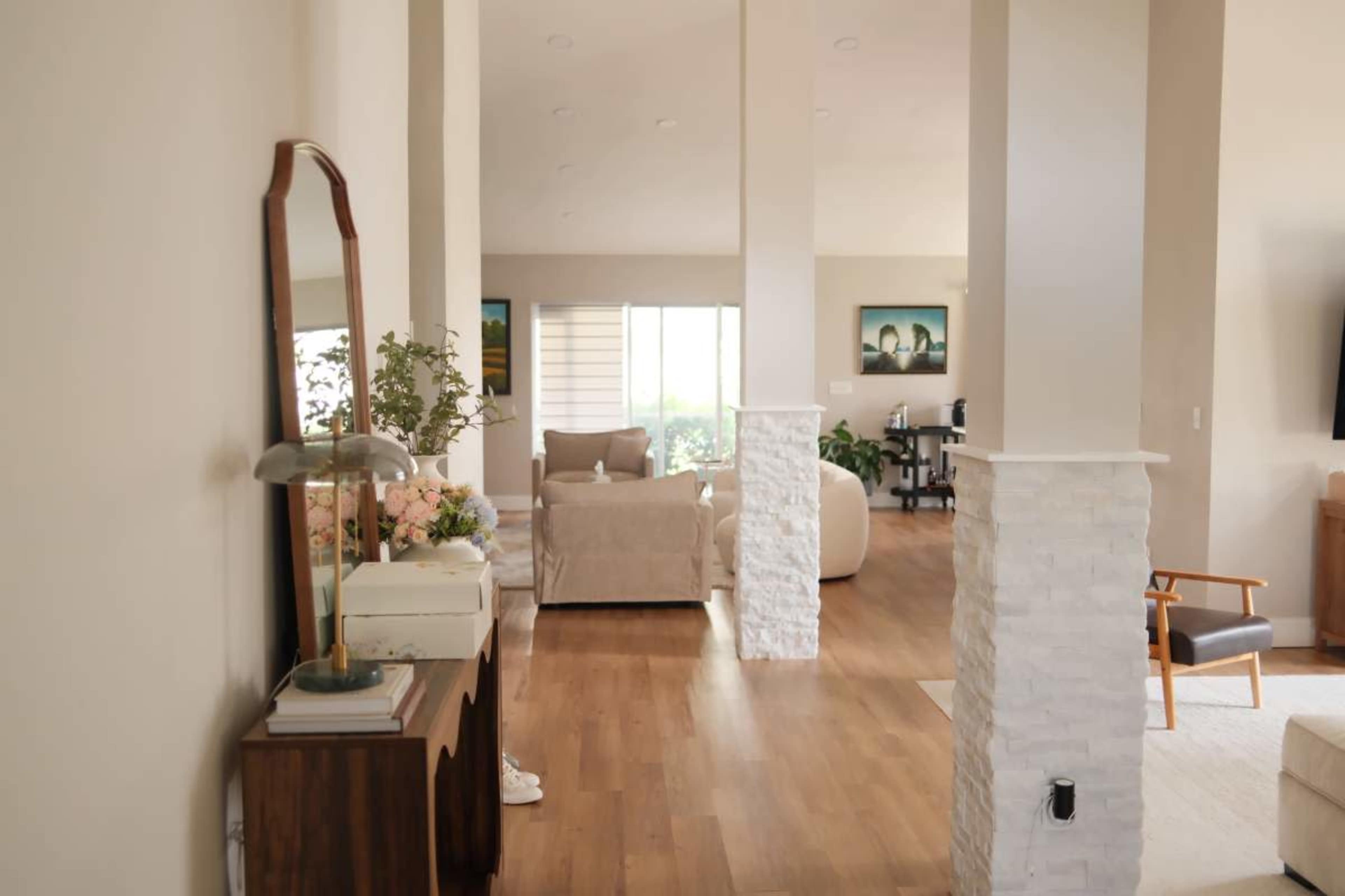 The image shows a spacious interior of a modern living area with light wooden flooring, a mix of furniture, and large windows allowing natural light to enter.