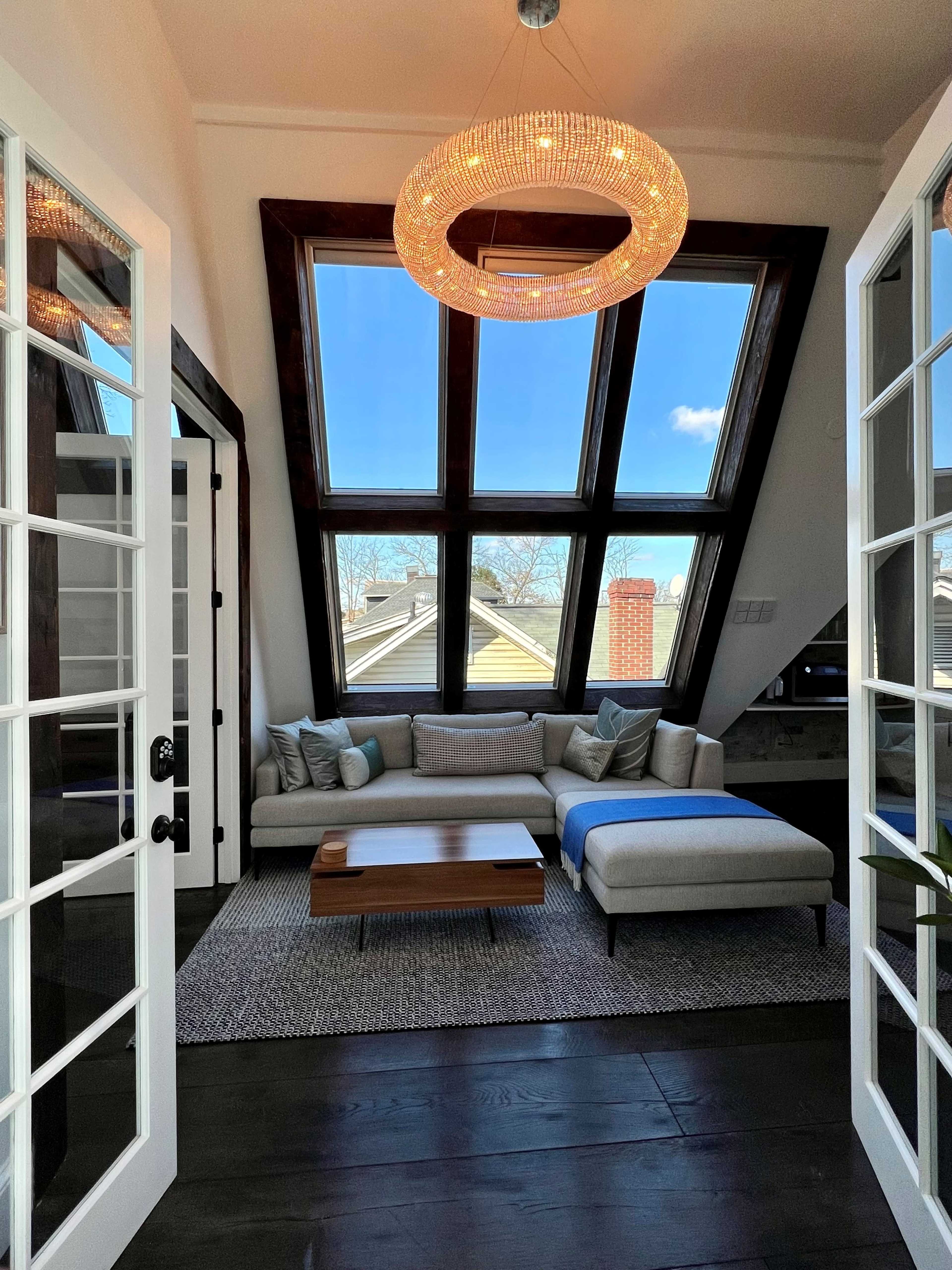 A cozy living area with large triangular windows, a light-colored sofa, a wooden coffee table, and a circular chandelier.