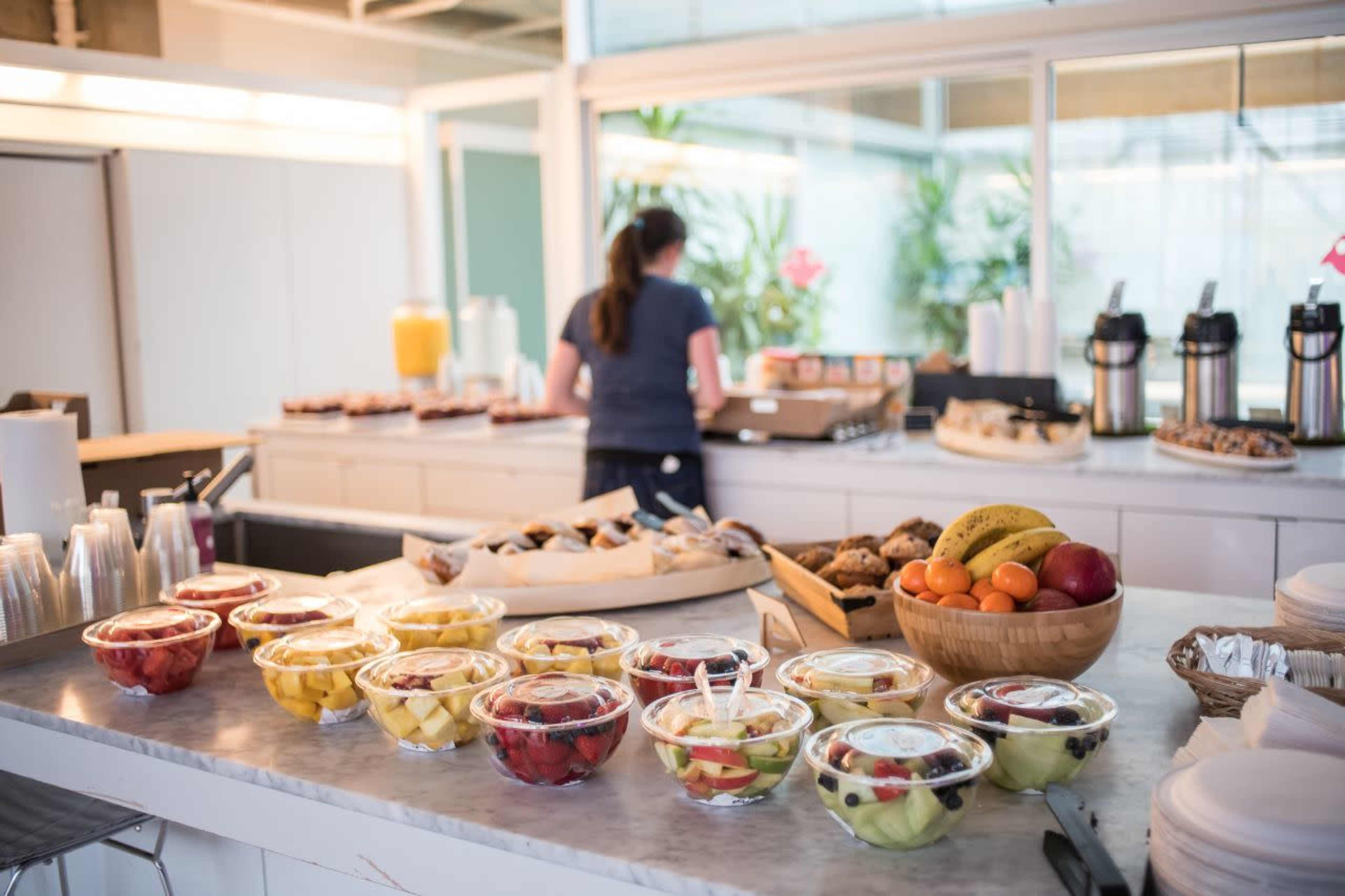 A buffet table displays an assortment of fruit cups and baked goods, while a person prepares food in the background.