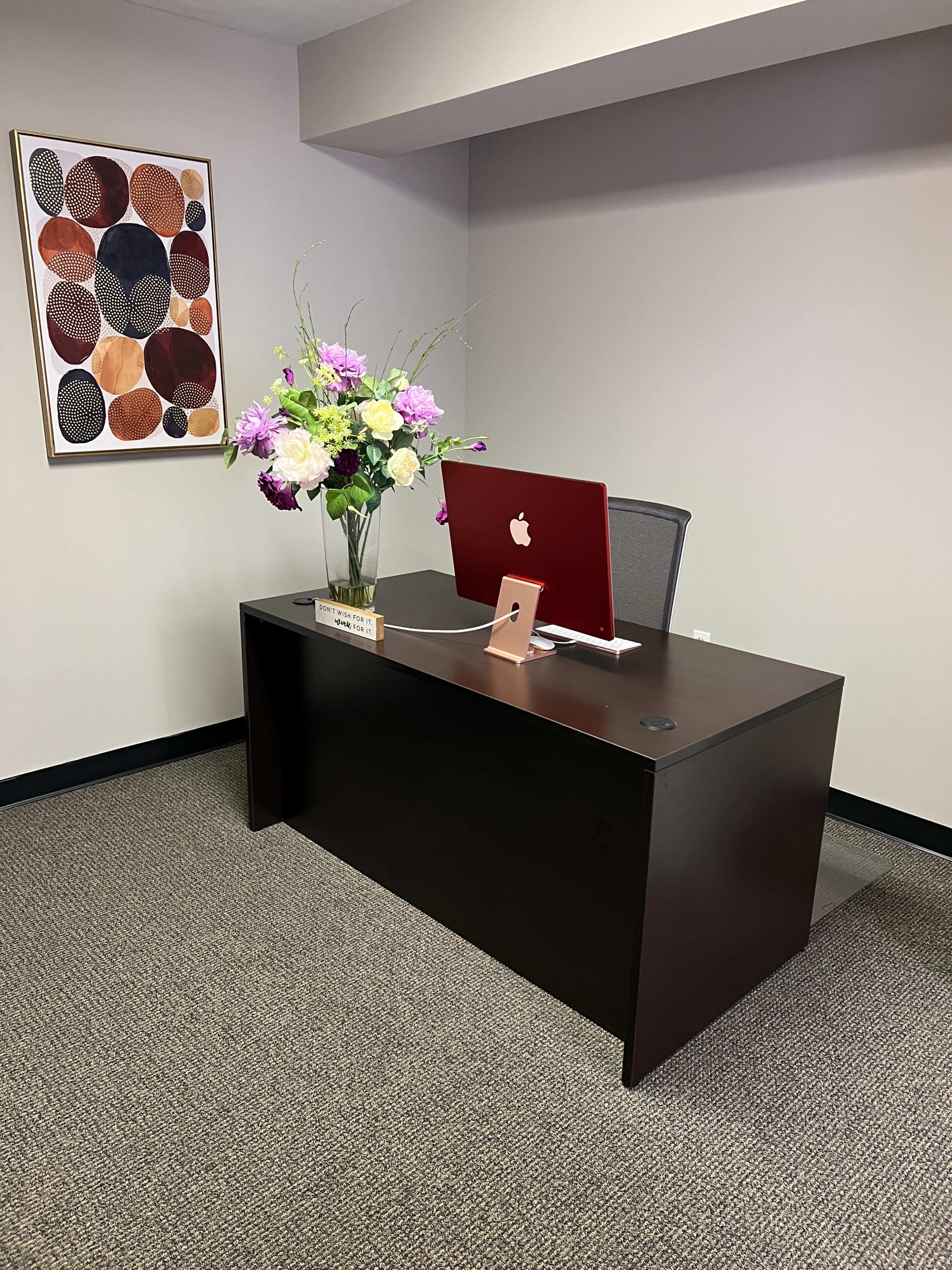 The image shows a modern office space featuring a dark wooden desk with an iMac computer, a decorative vase of flowers, and framed artwork on the wall.