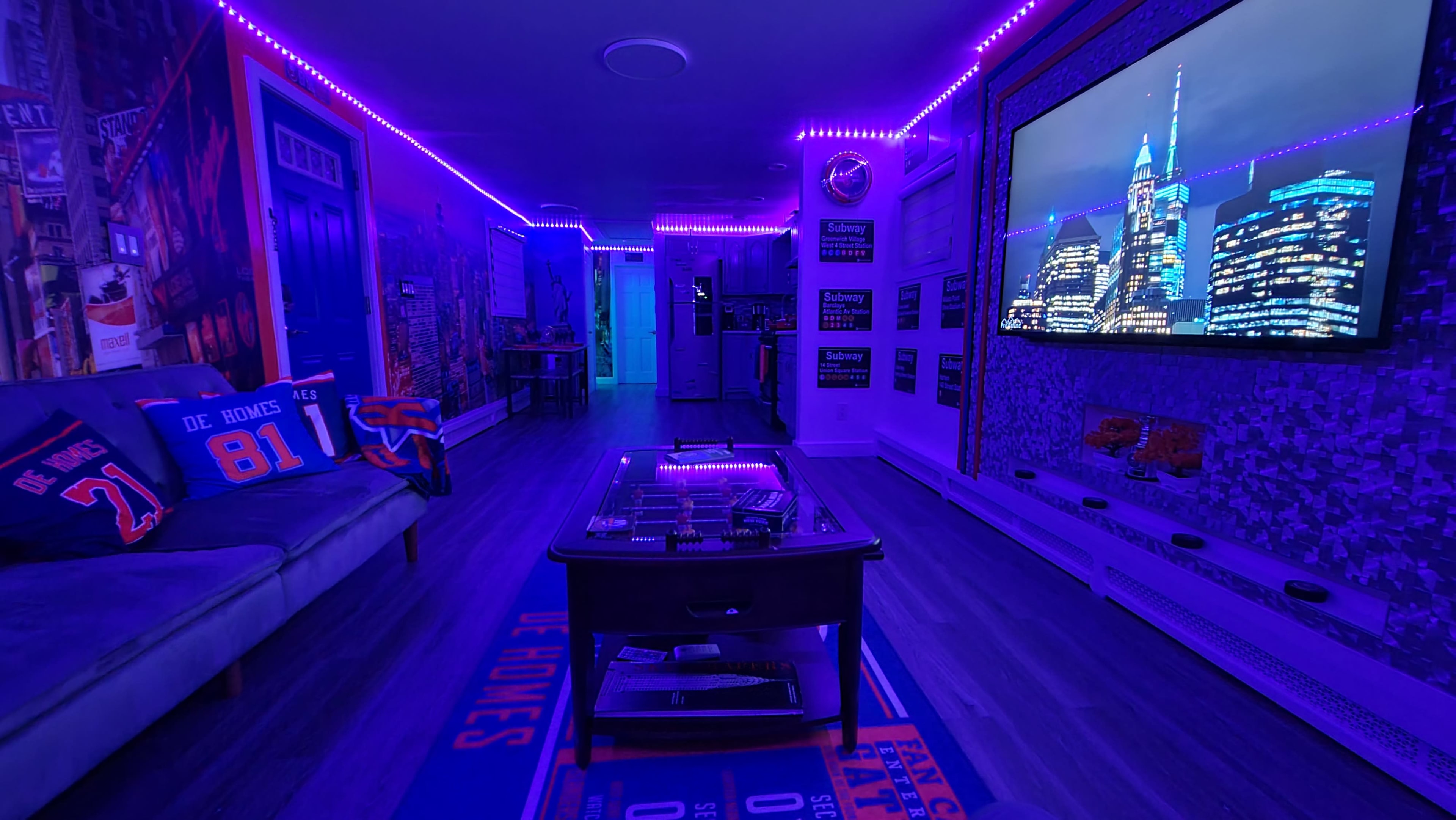 The room features a modern living space with purple LED lights, a couch, a coffee table, and a large screen displaying a city skyline.