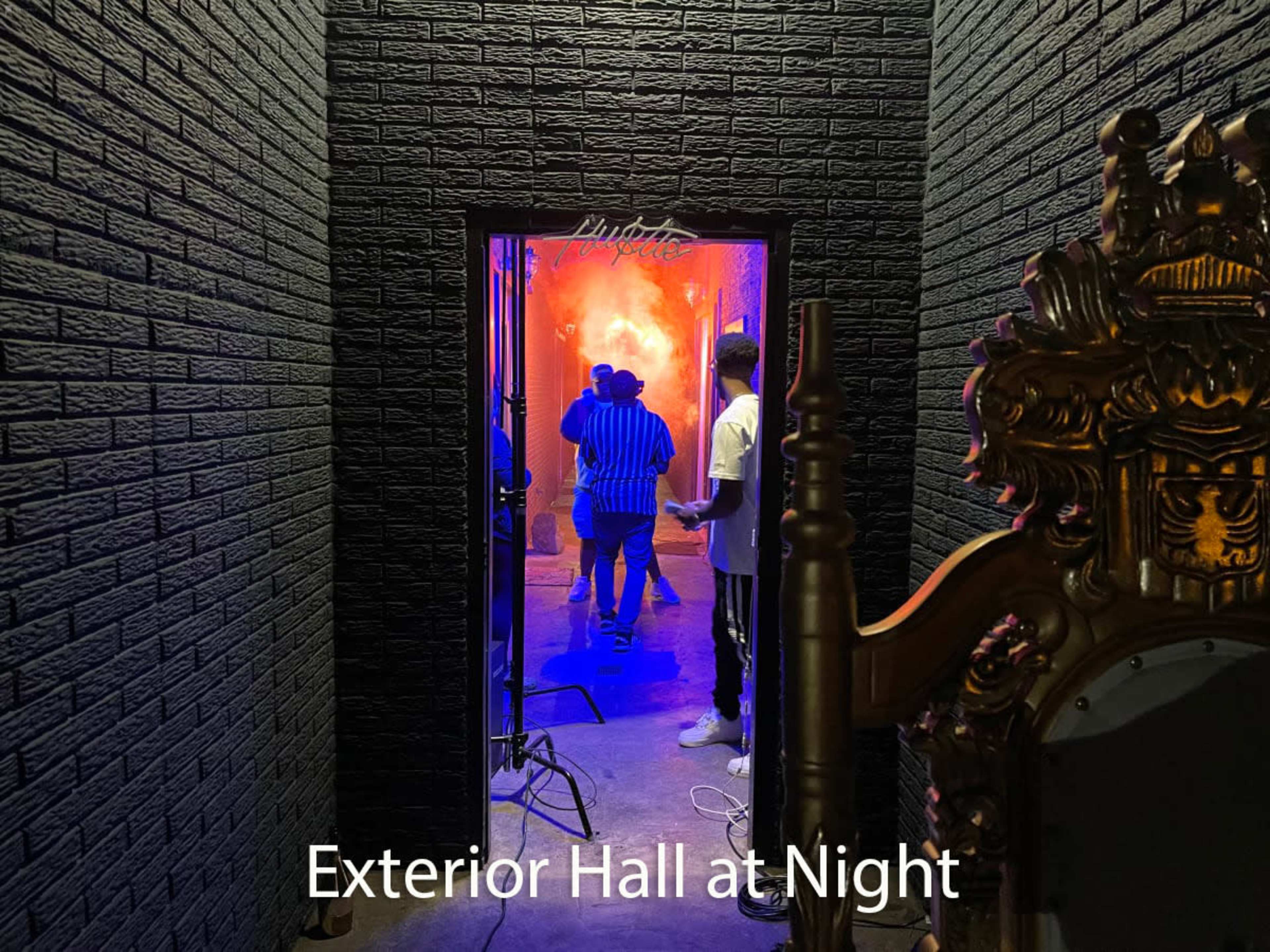 The image shows a dimly lit exterior hall at night, with a doorway revealing a lit space and several people visible inside.