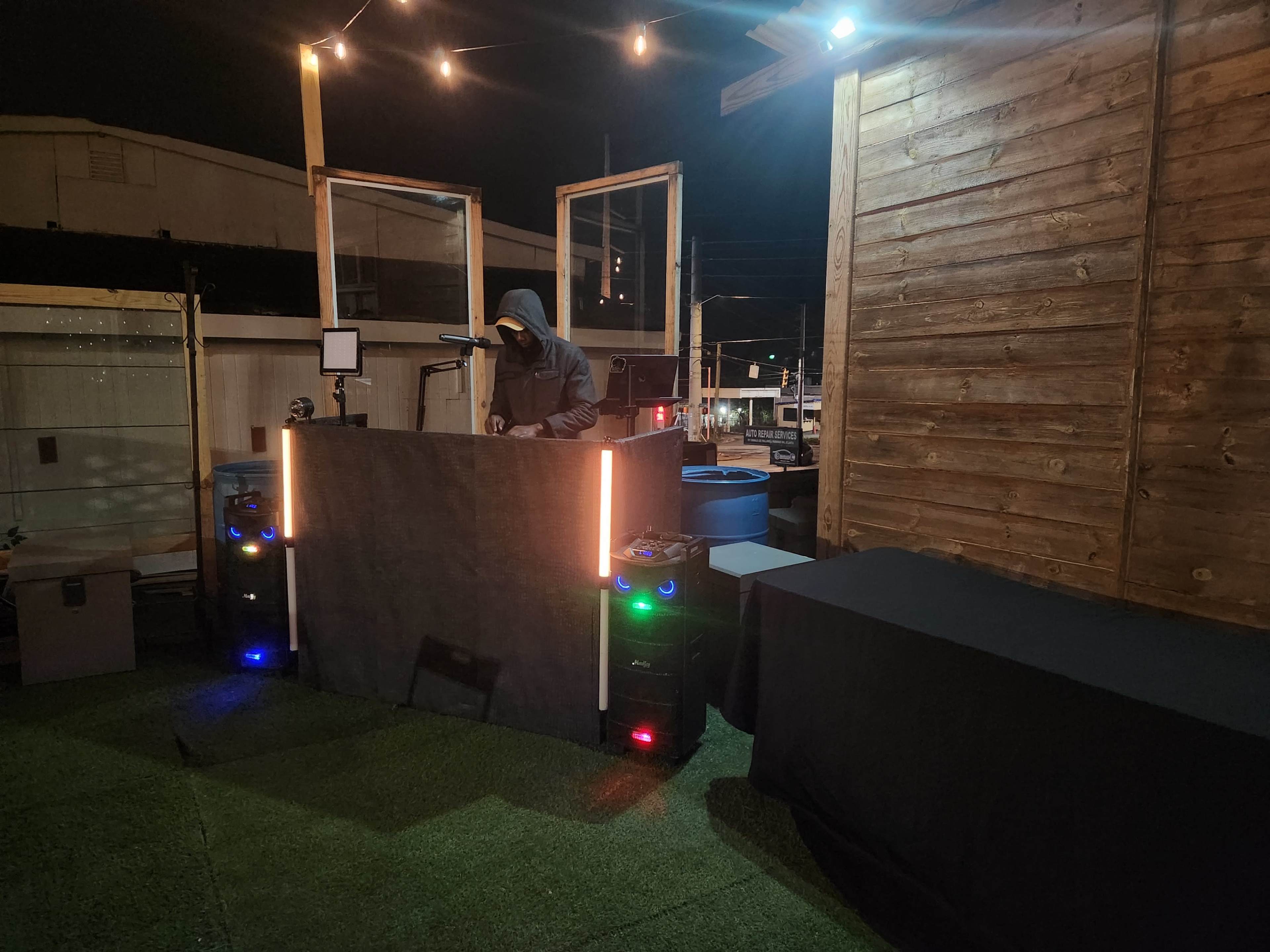A DJ, dressed in a hooded sweatshirt, stands behind a dark, illuminated booth on an outdoor deck, with speakers beside him and string lights overhead.