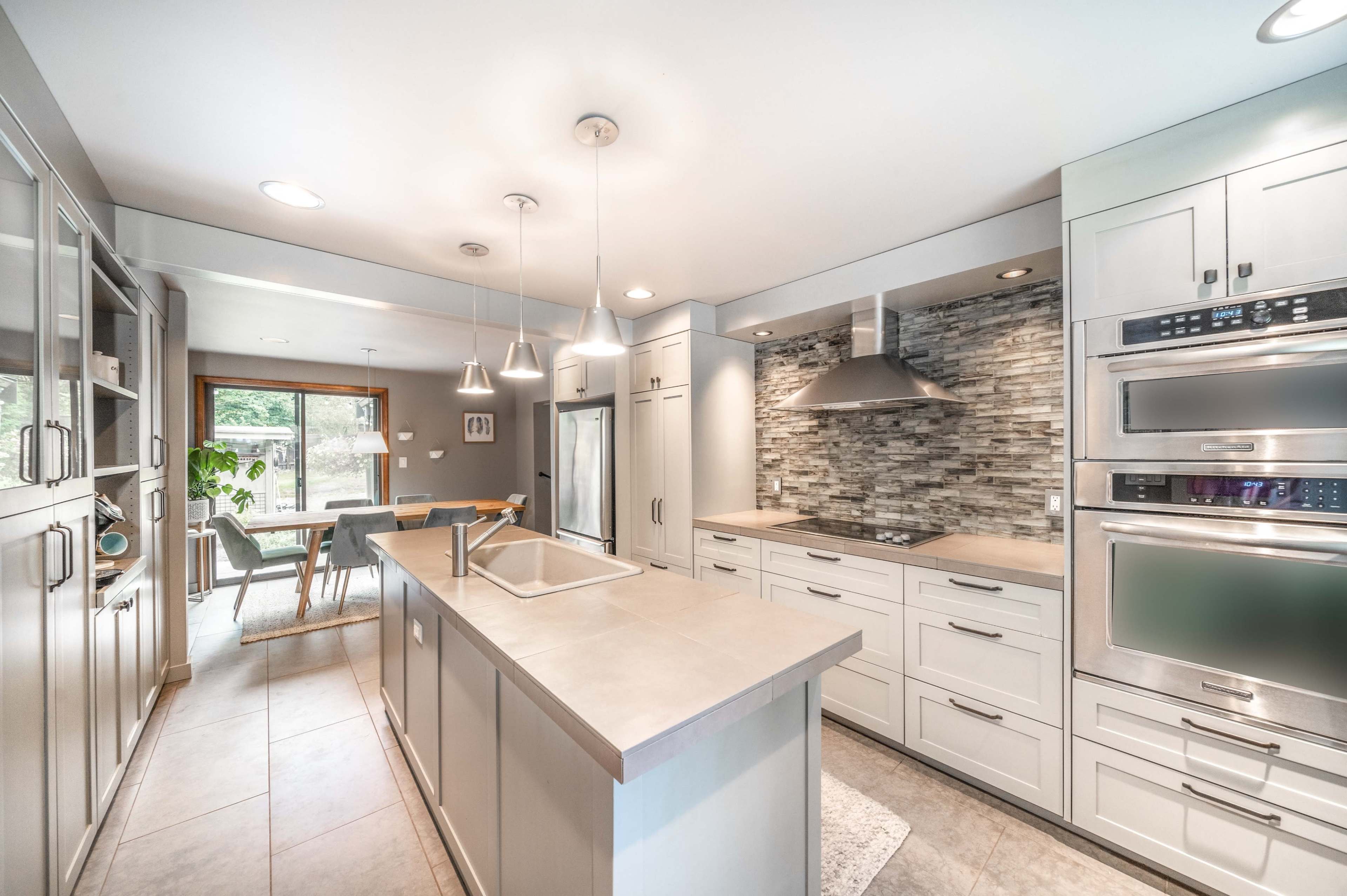 The image shows a modern kitchen with light cabinetry, a central island, and stainless steel appliances, featuring a textured stone backsplash.