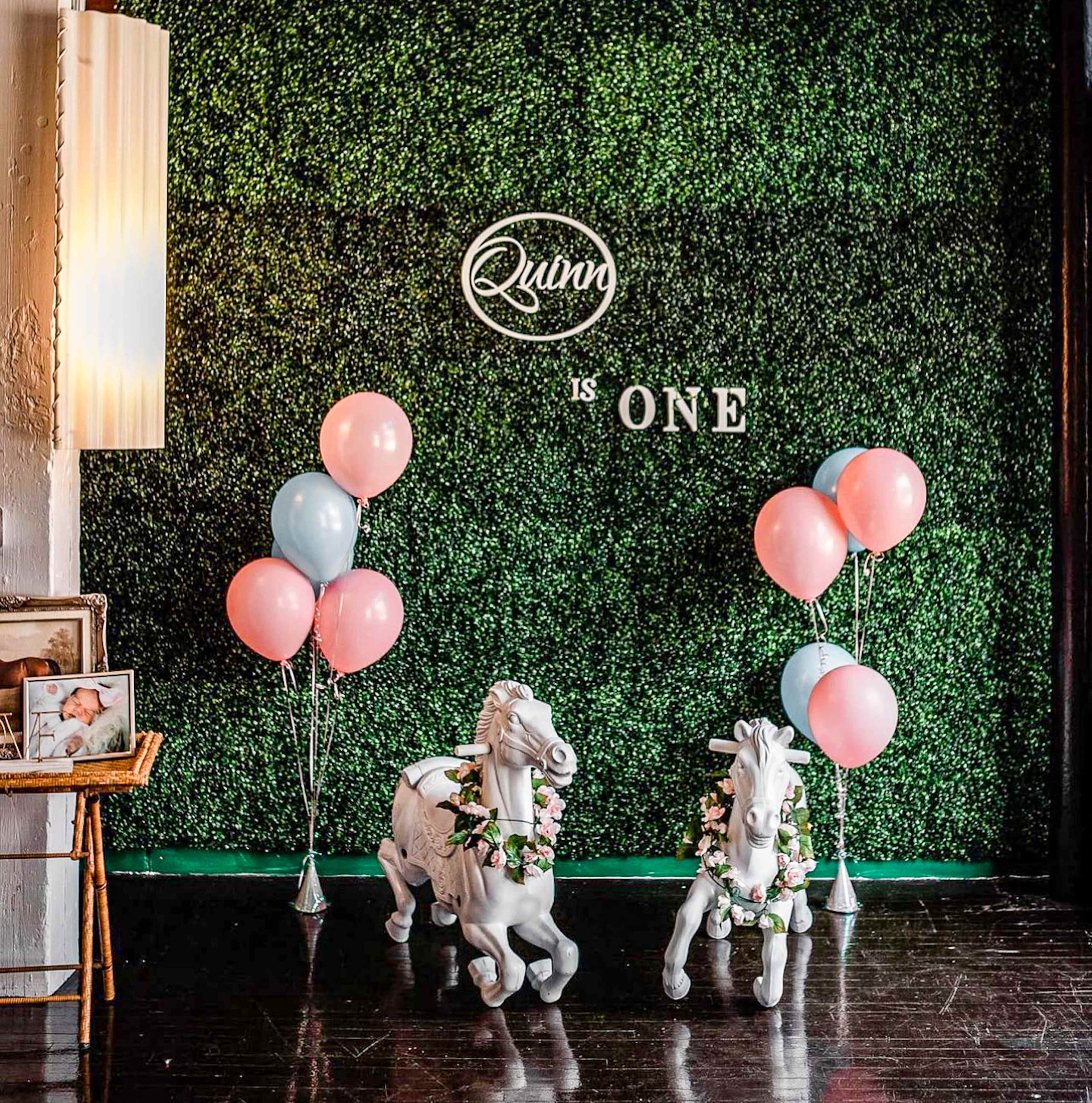 The scene features a green wall adorned with the words "Quinn is ONE," complemented by two white toy horses decorated with flowers and pastel pink and blue balloons.