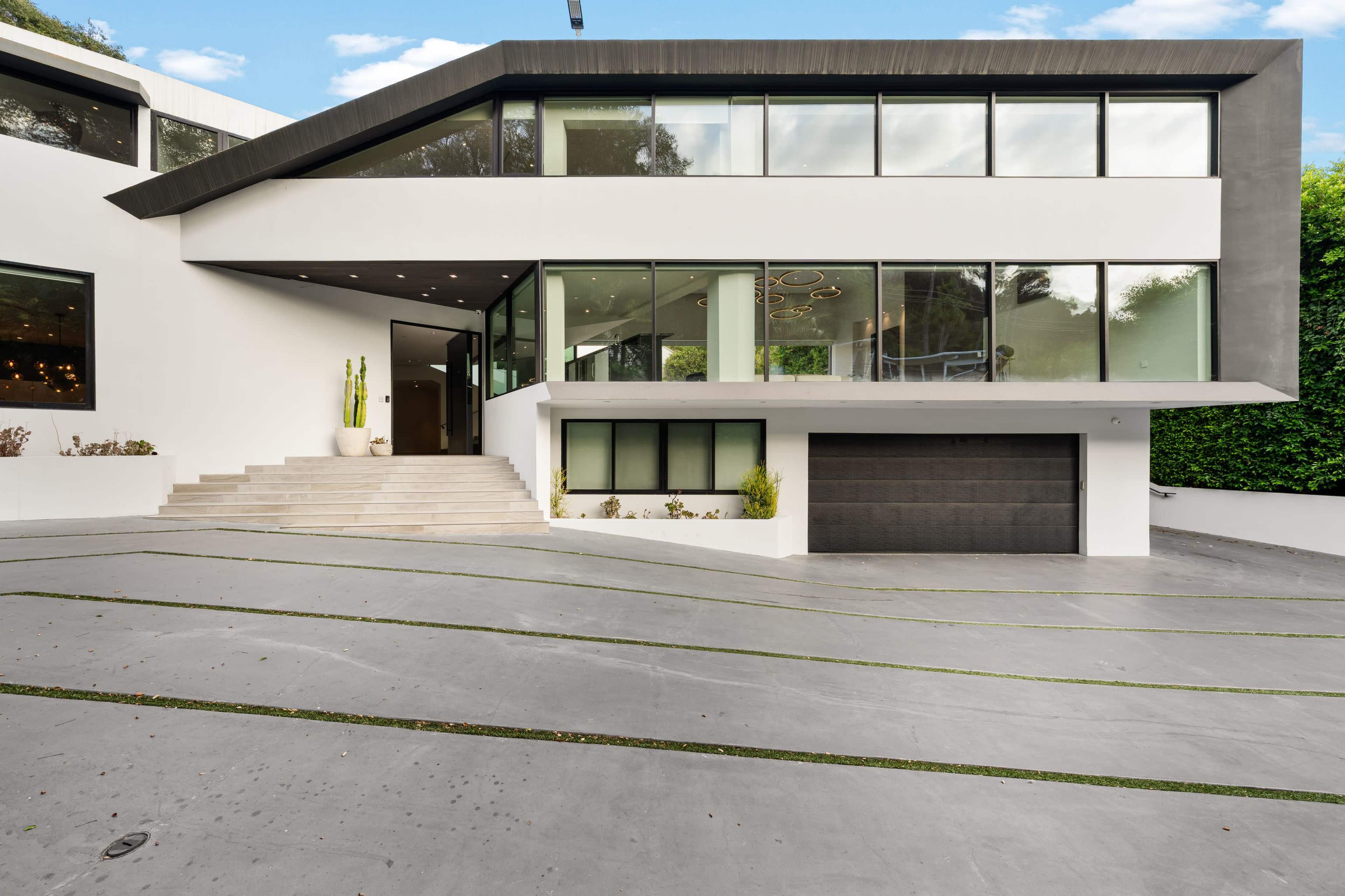 The image shows a modern architectural house featuring large glass windows, a stylish entrance, and a driveway leading to a garage.