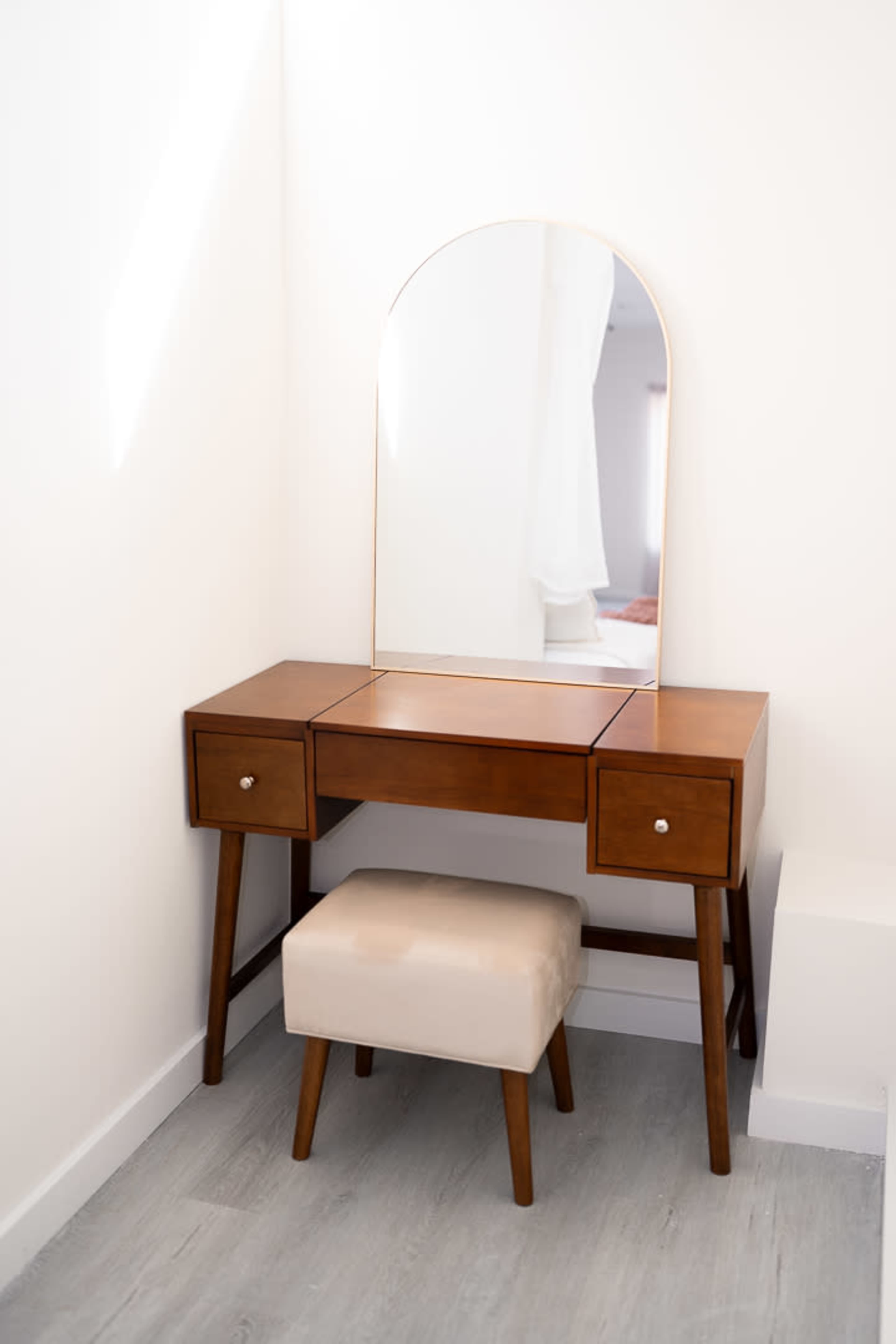 A wooden vanity table with a large arched mirror and a beige upholstered stool is situated in a bright, minimalist corner.