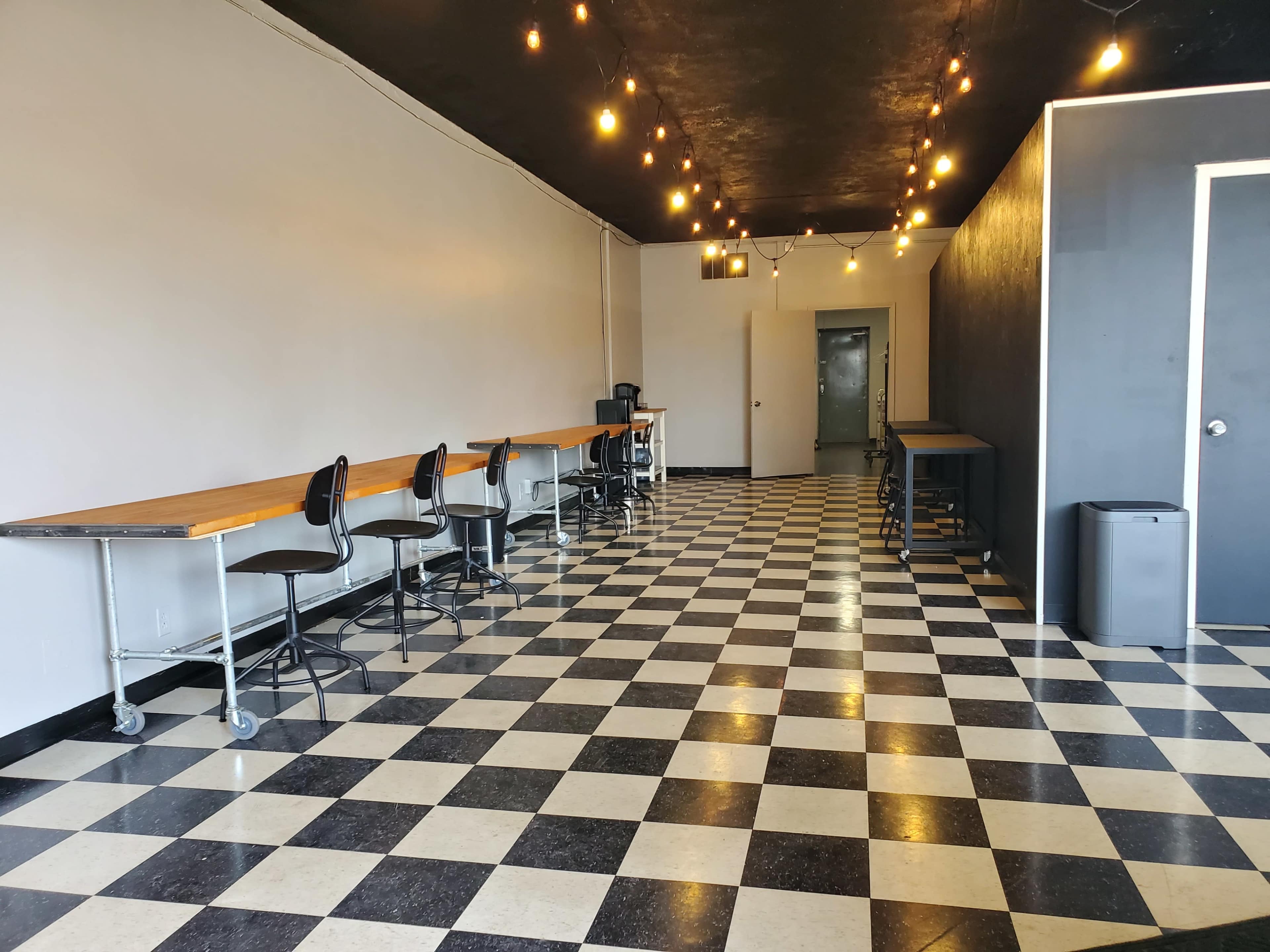 A spacious interior with black and white checkered flooring, featuring several bar-height stools at long tables and pendant lights hanging from the ceiling.