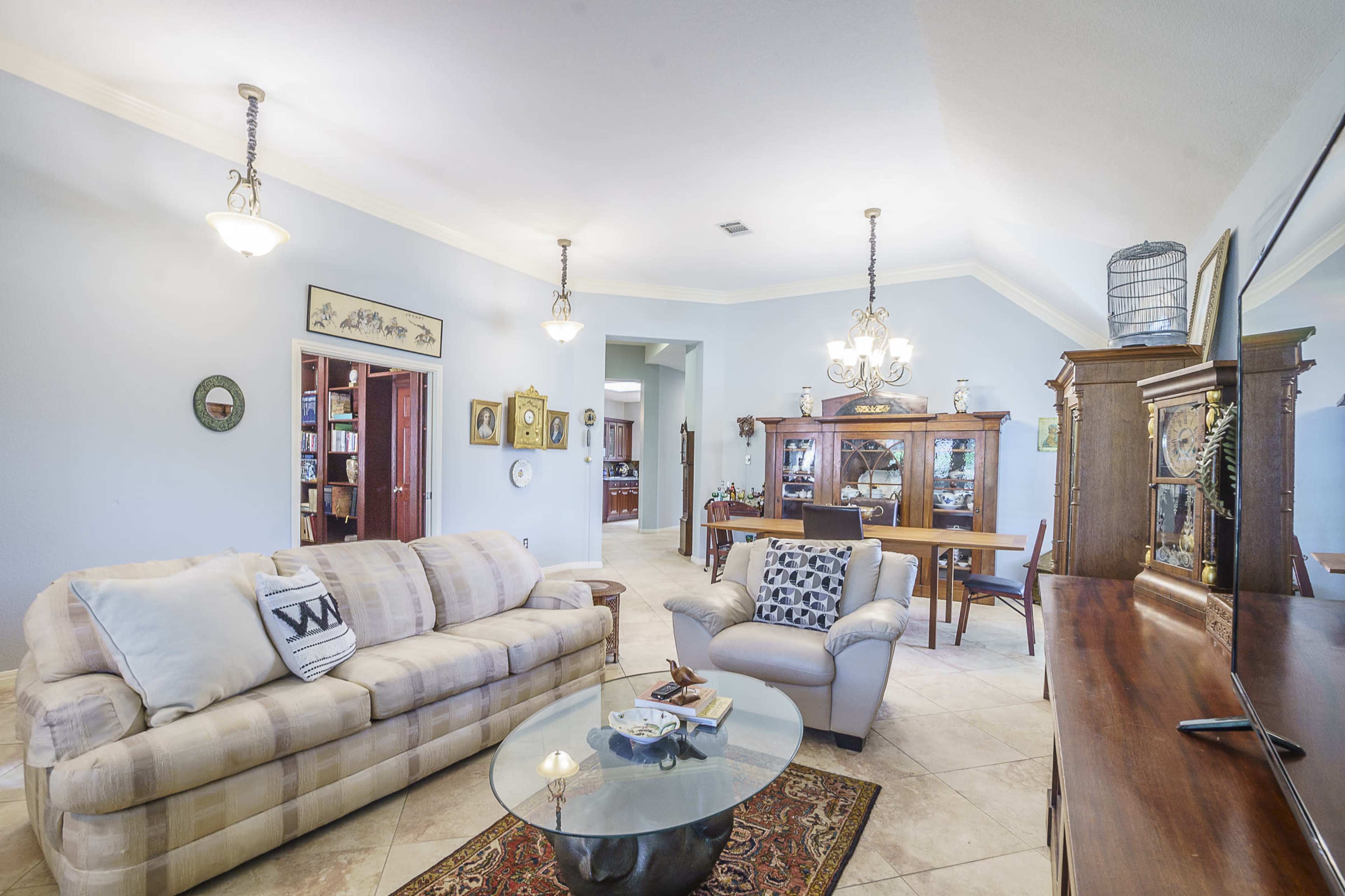 A spacious living room features a light-colored sofa and armchair arranged around a glass coffee table, with a dining area and wooden furniture visible in the background.