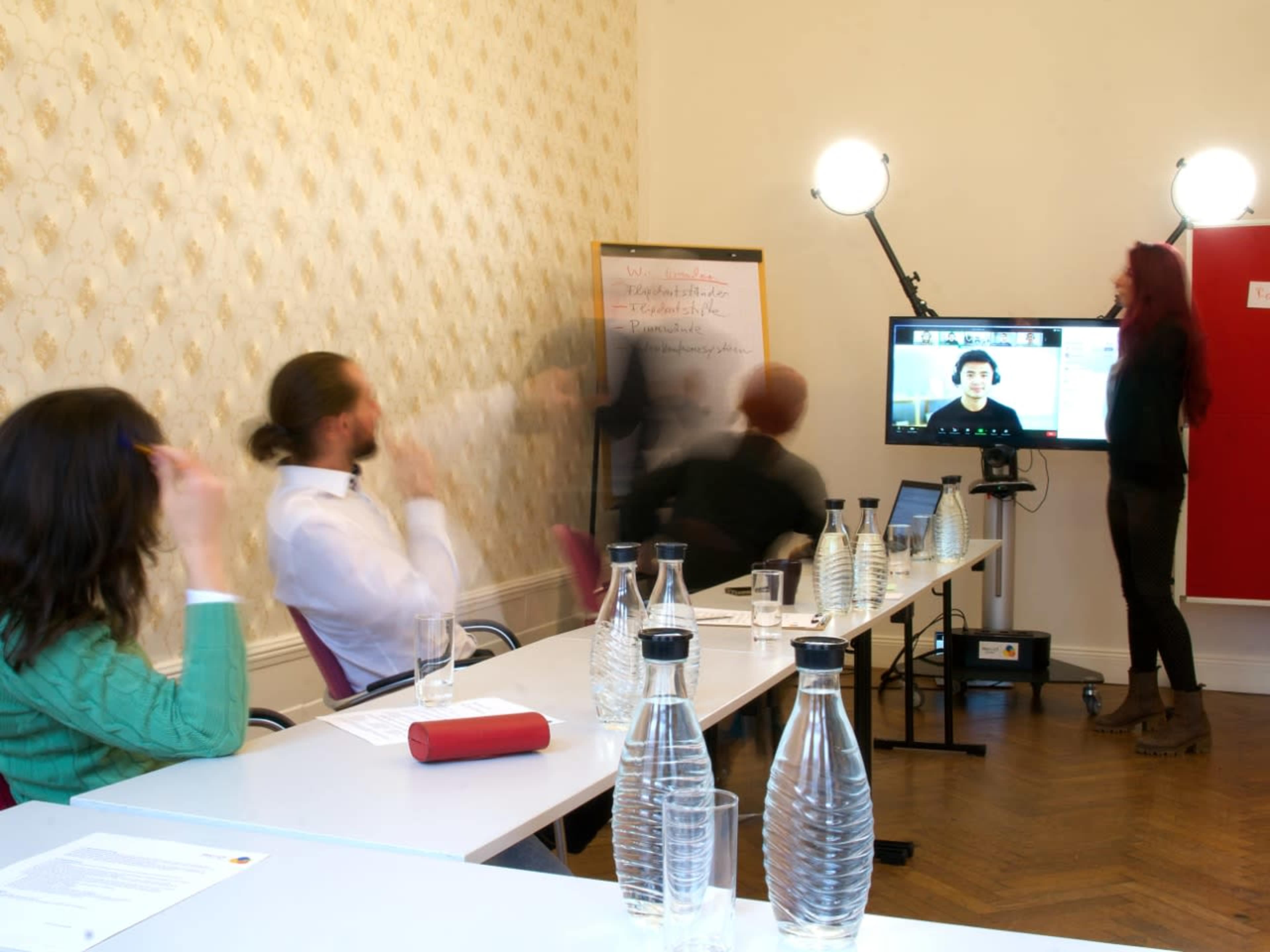 A group of people participates in a meeting, with some attendees on a video call displayed on a screen while others engage in discussion at a table with water bottles.