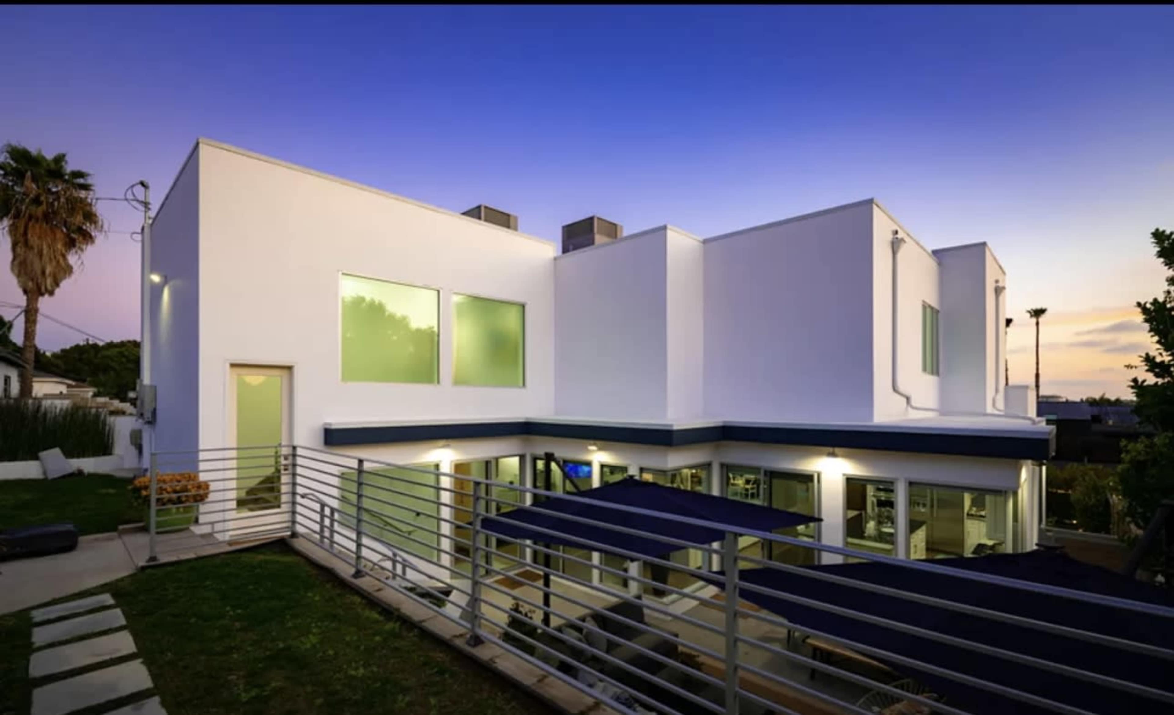 A modern white house with large windows and a sleek design is surrounded by greenery under a dusk sky.