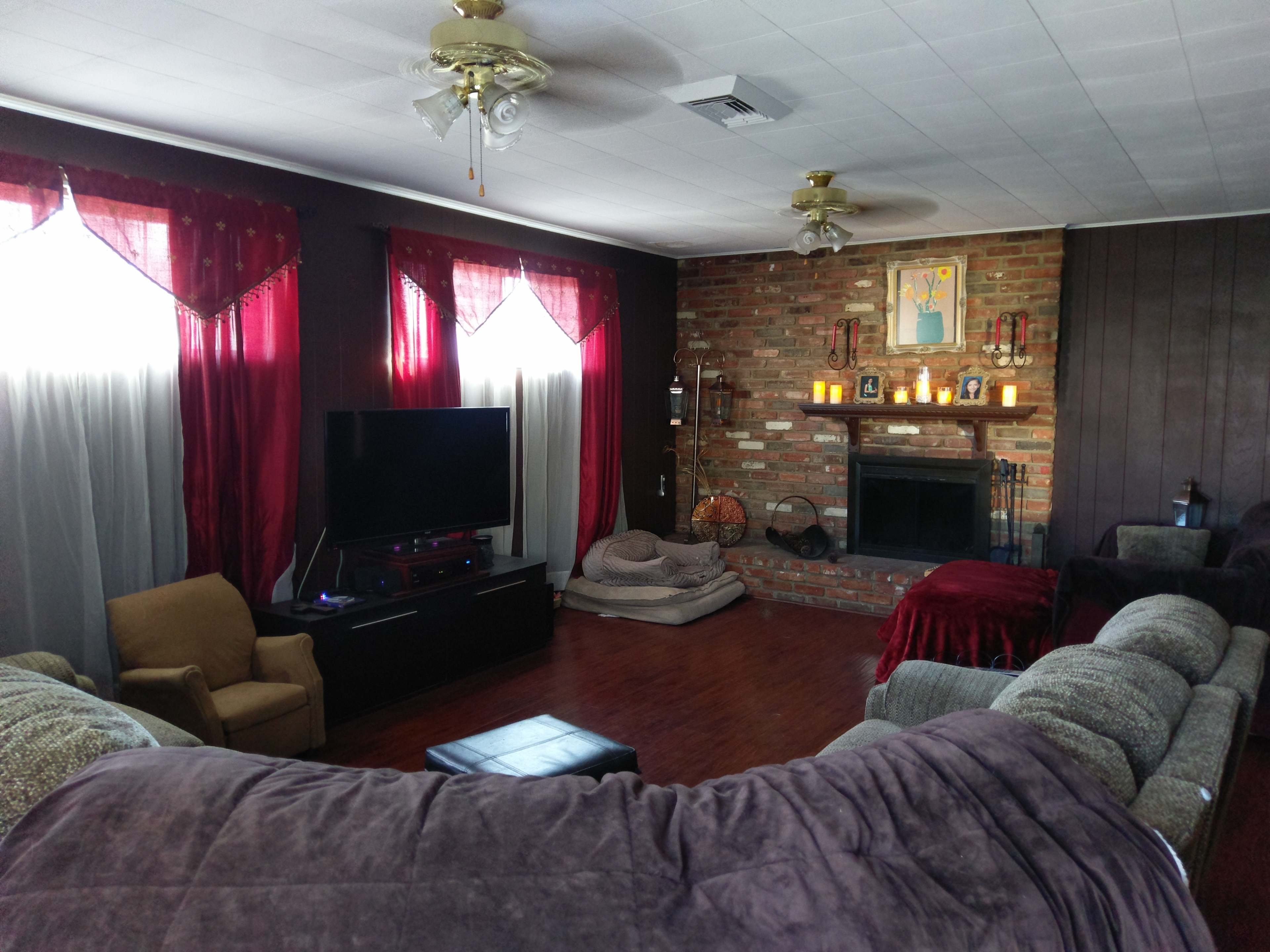 The living room features a sectional sofa, a television unit, a fireplace with candles, and large windows adorned with red curtains.