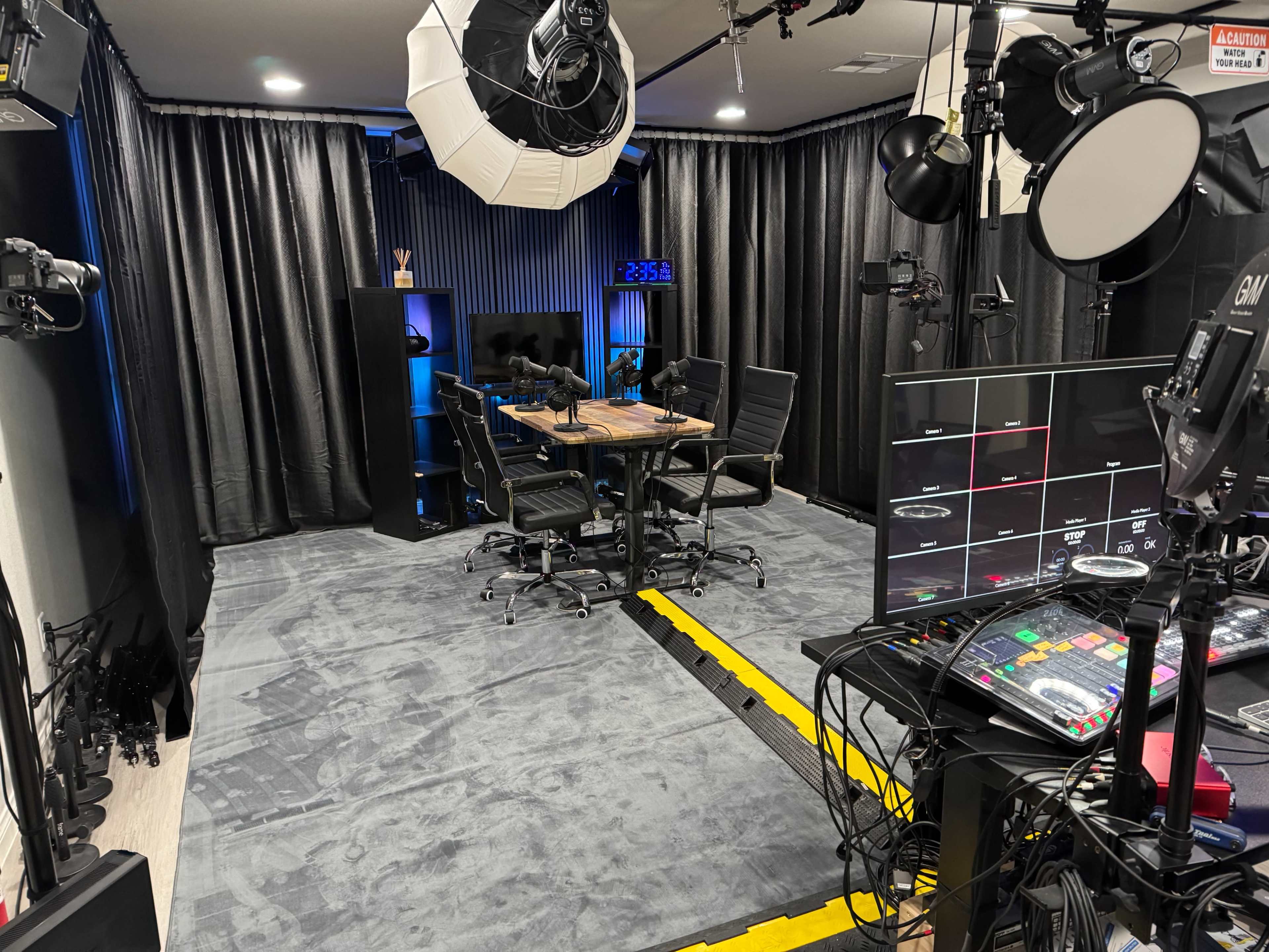 The image shows a well-equipped multimedia studio featuring several cameras, lighting equipment, and a control station with monitors and a mixer, all arranged around a central table with chairs.