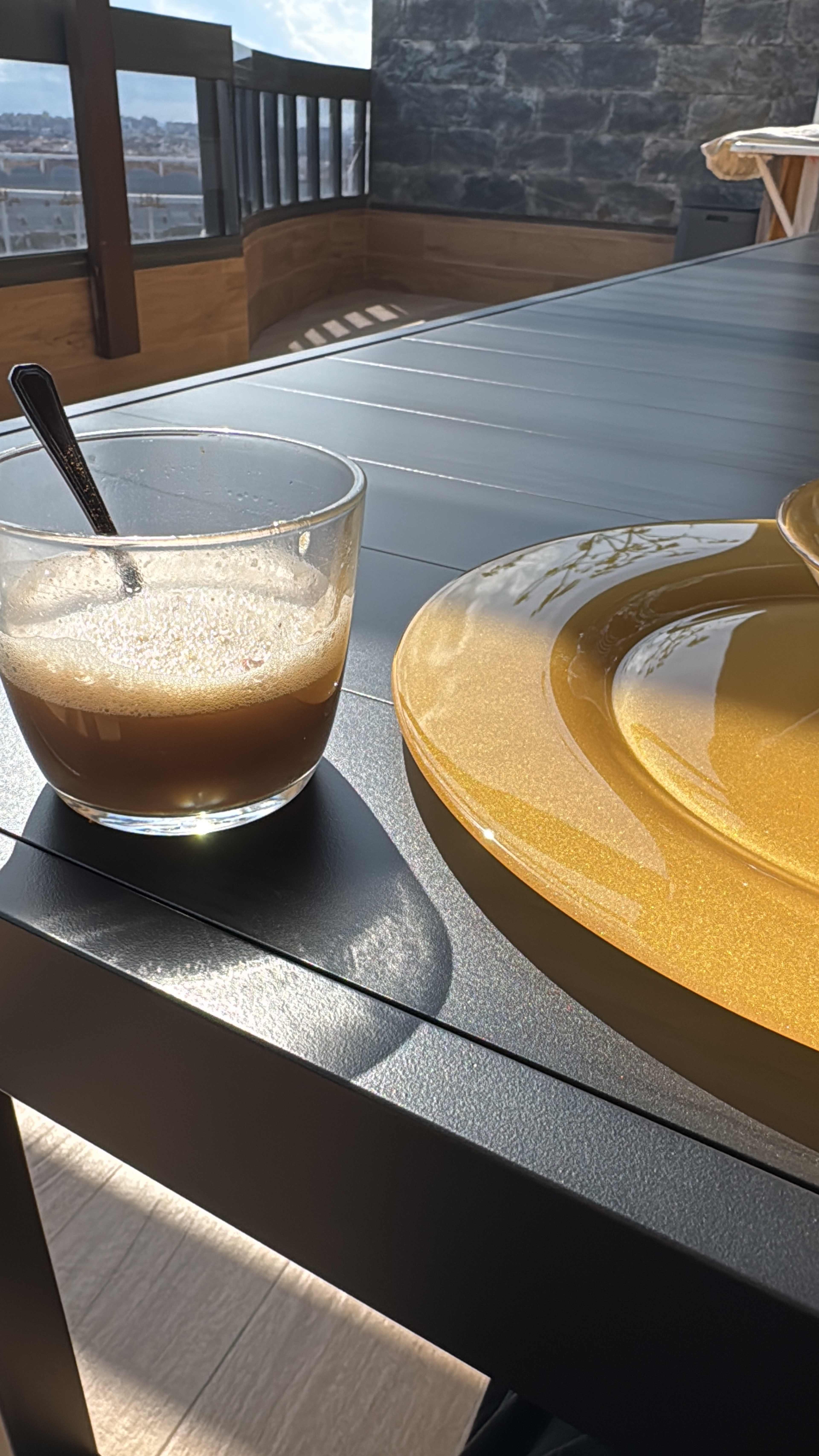 A glass of coffee sits next to a yellow plate on a wooden table with a view in the background.