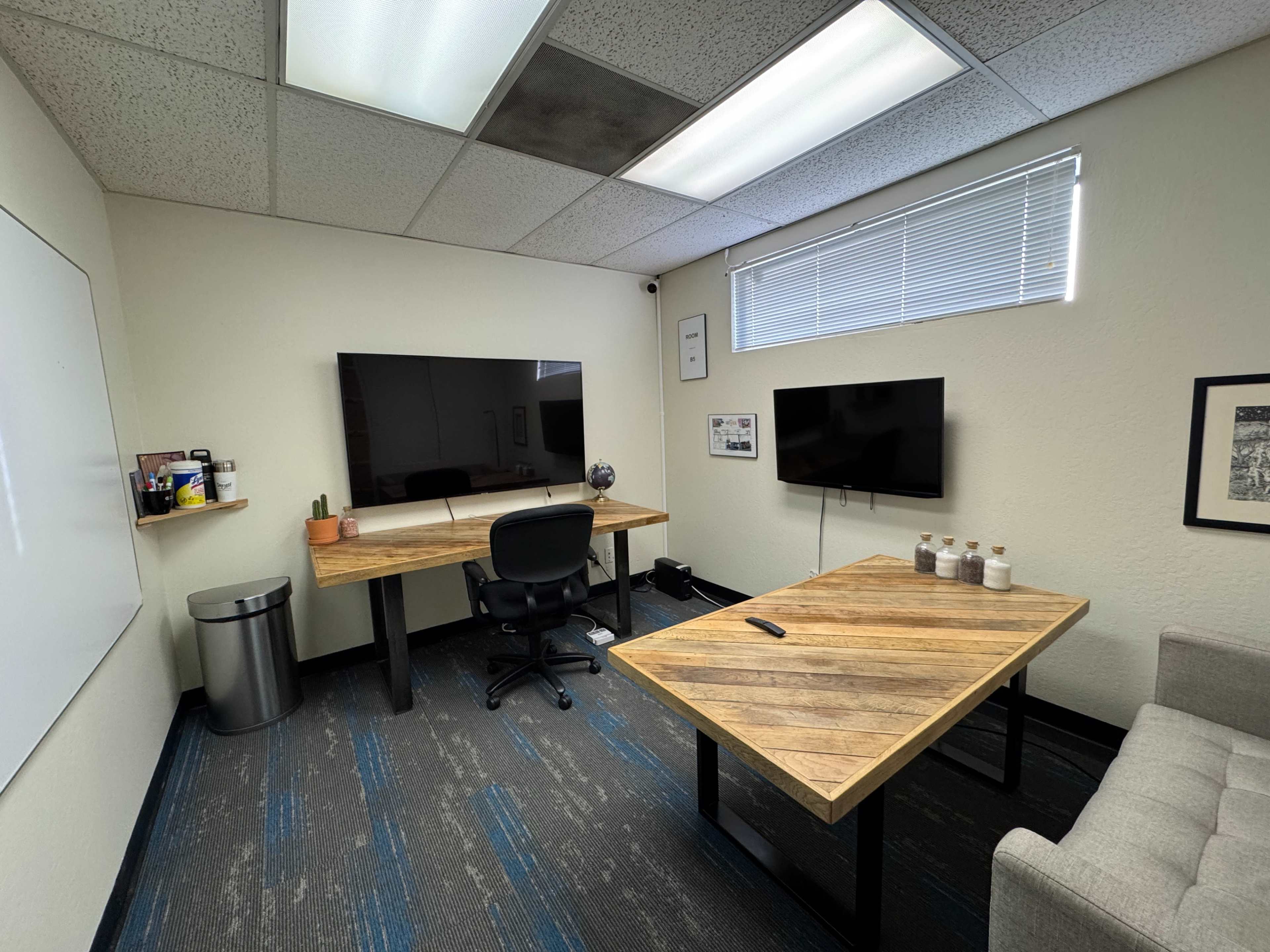 The image shows a small office space featuring two wooden desks, two wall-mounted TVs, a chair, a waste bin, and a gray couch.