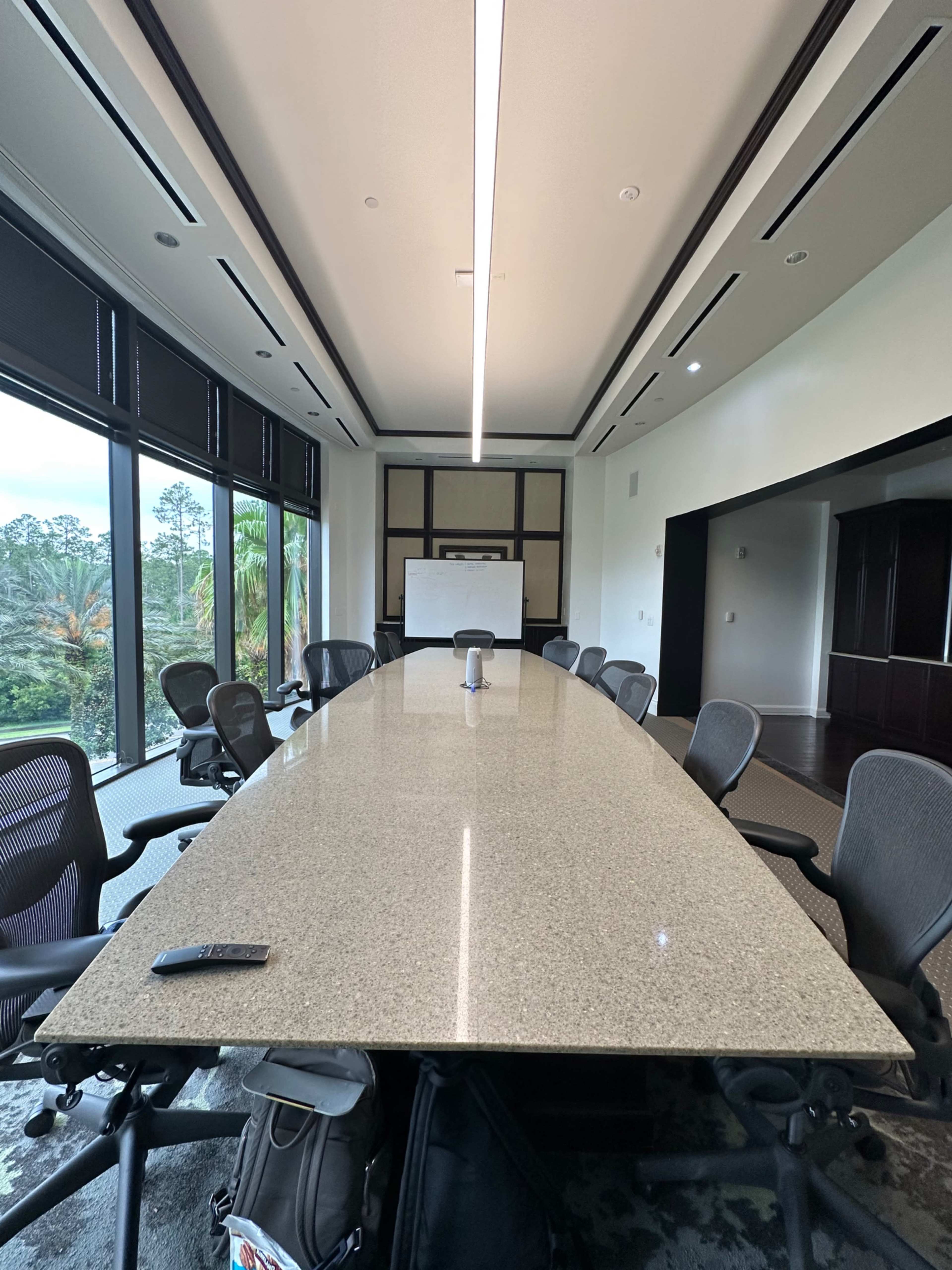 A large, modern conference room with a long, smooth table surrounded by office chairs and large windows overlooking a green landscape.