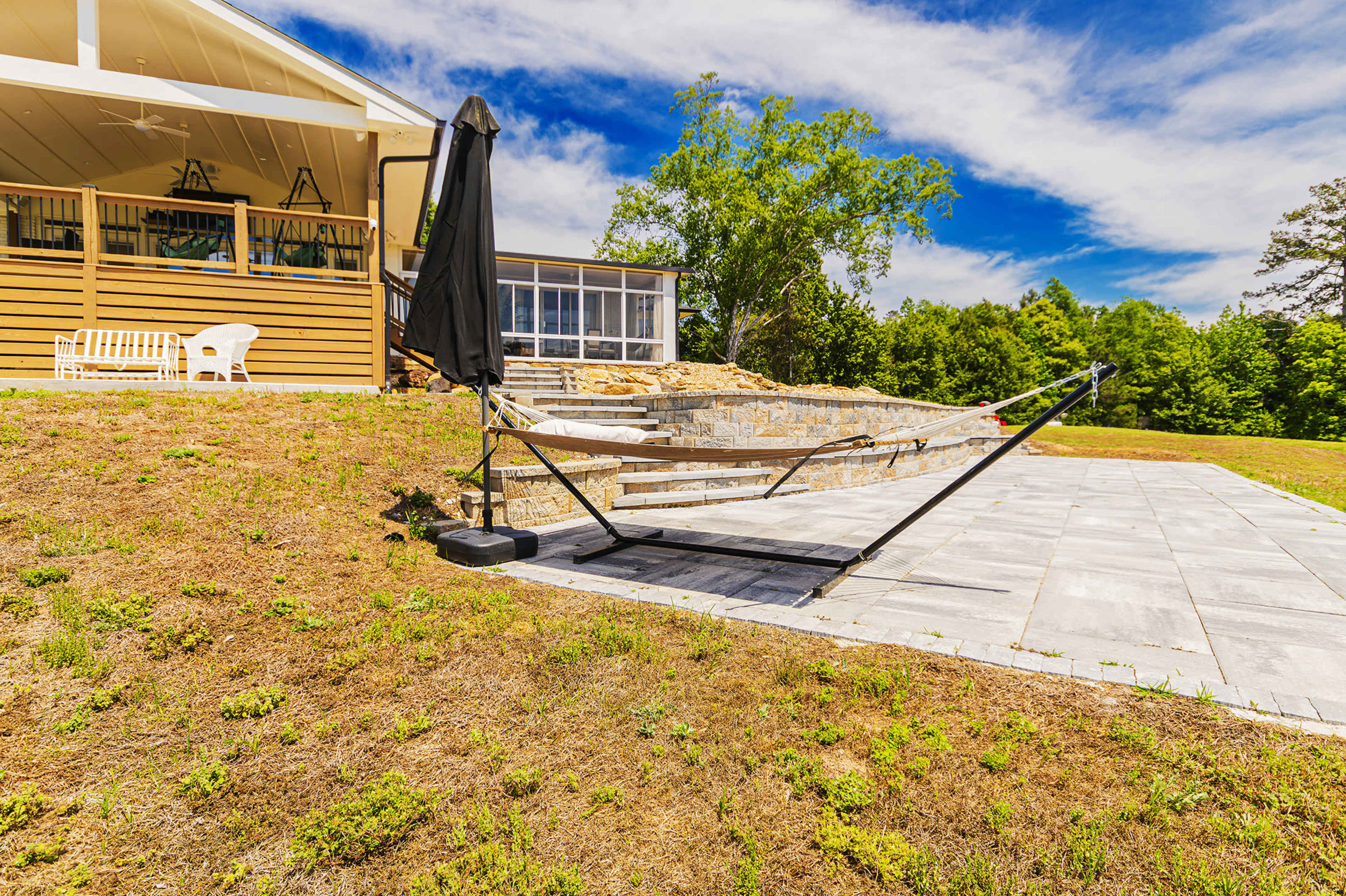 A hammock stand with a black umbrella is positioned on a stone patio beside a house, with a grassy area behind it and trees in the background.