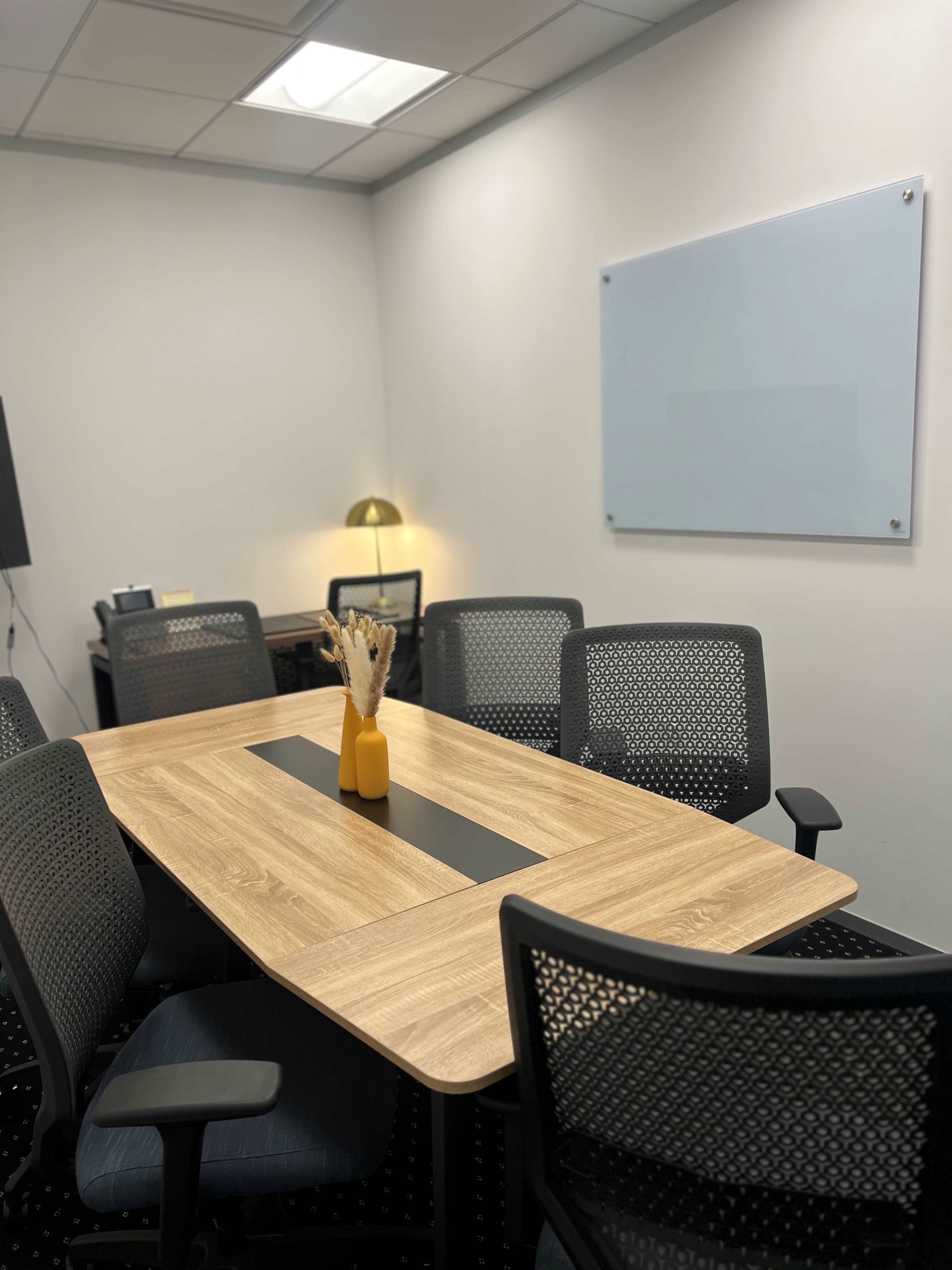 A modern conference room features a wooden table surrounded by mesh chairs, with a small vase of dried flowers in the center and a whiteboard mounted on the wall.