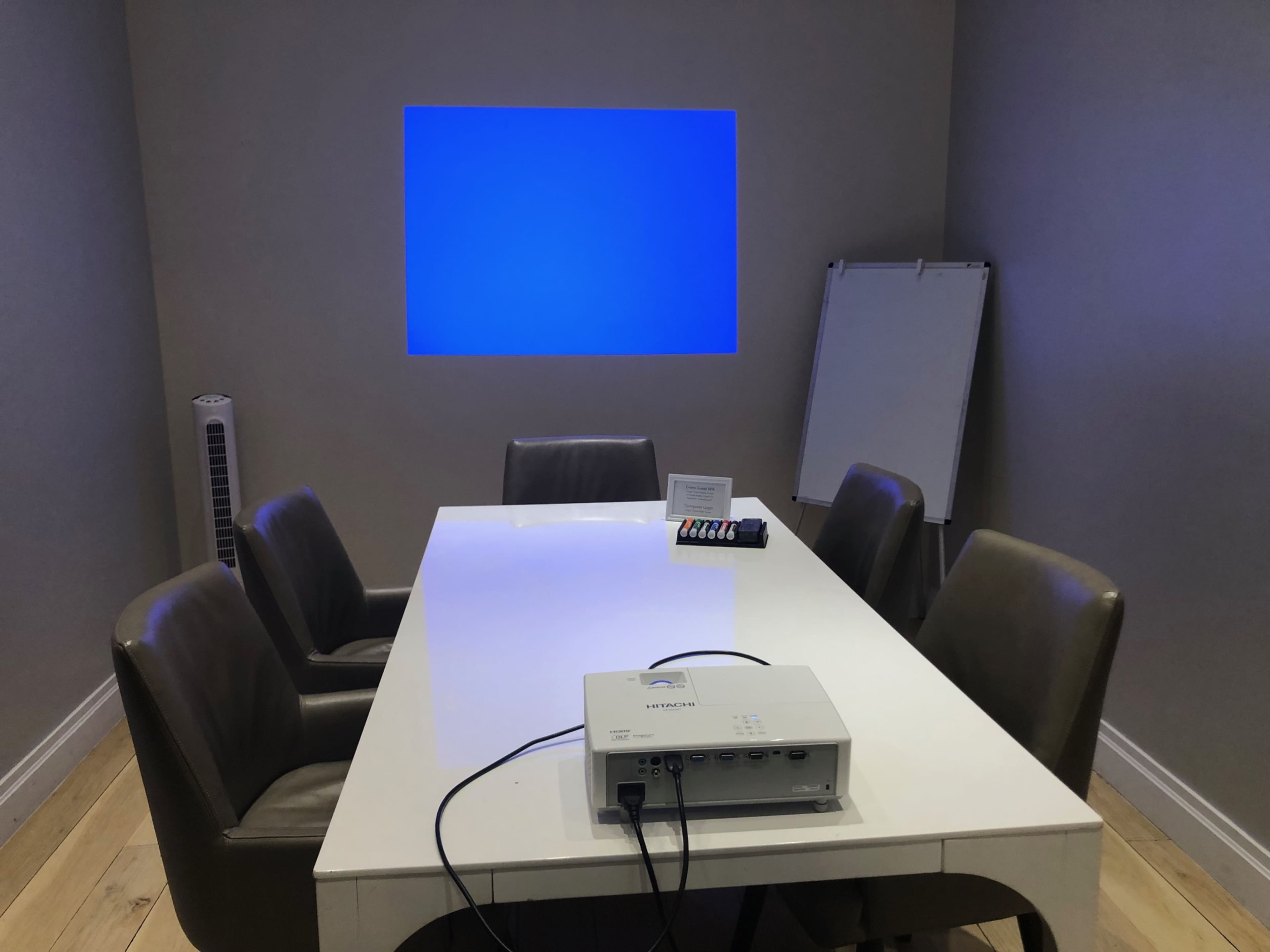 A conference room with a large white table, four chairs, a projector on the table, and a blue projection on the wall.