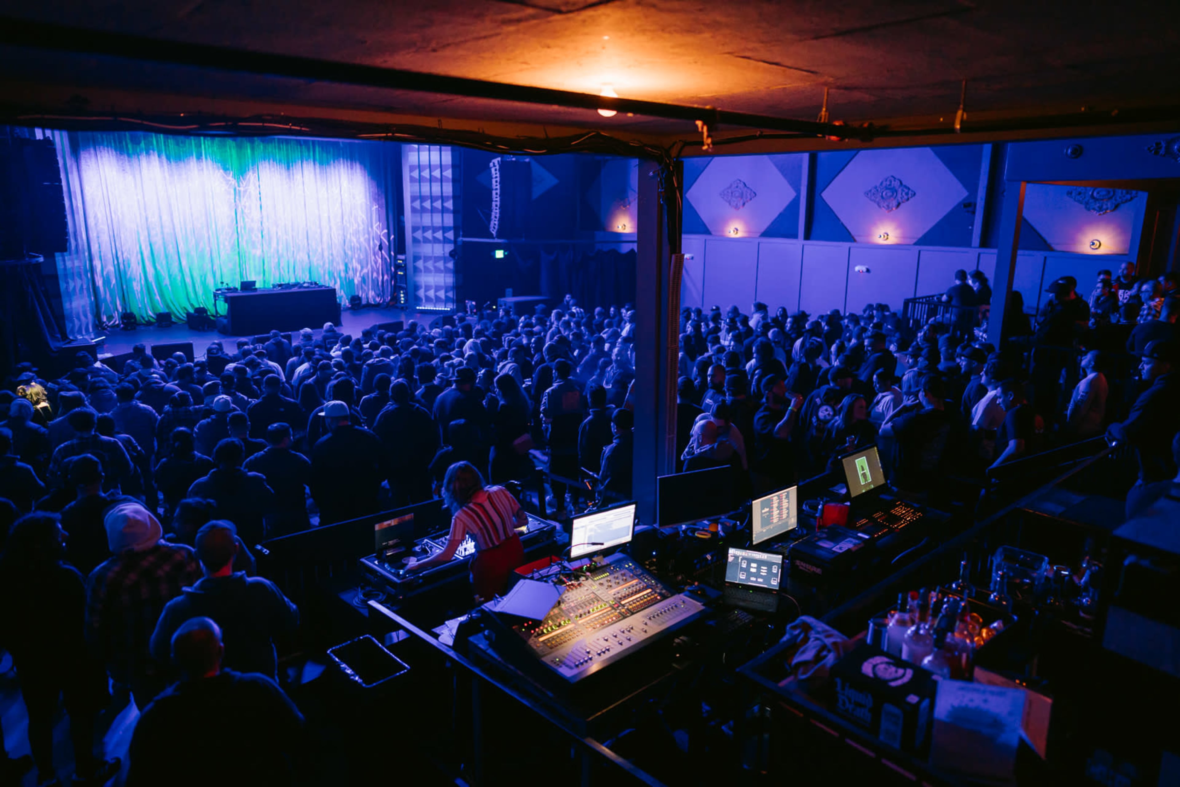 A crowded venue with a stage and lighting setup prepares for a performance, viewed from an elevated angle.