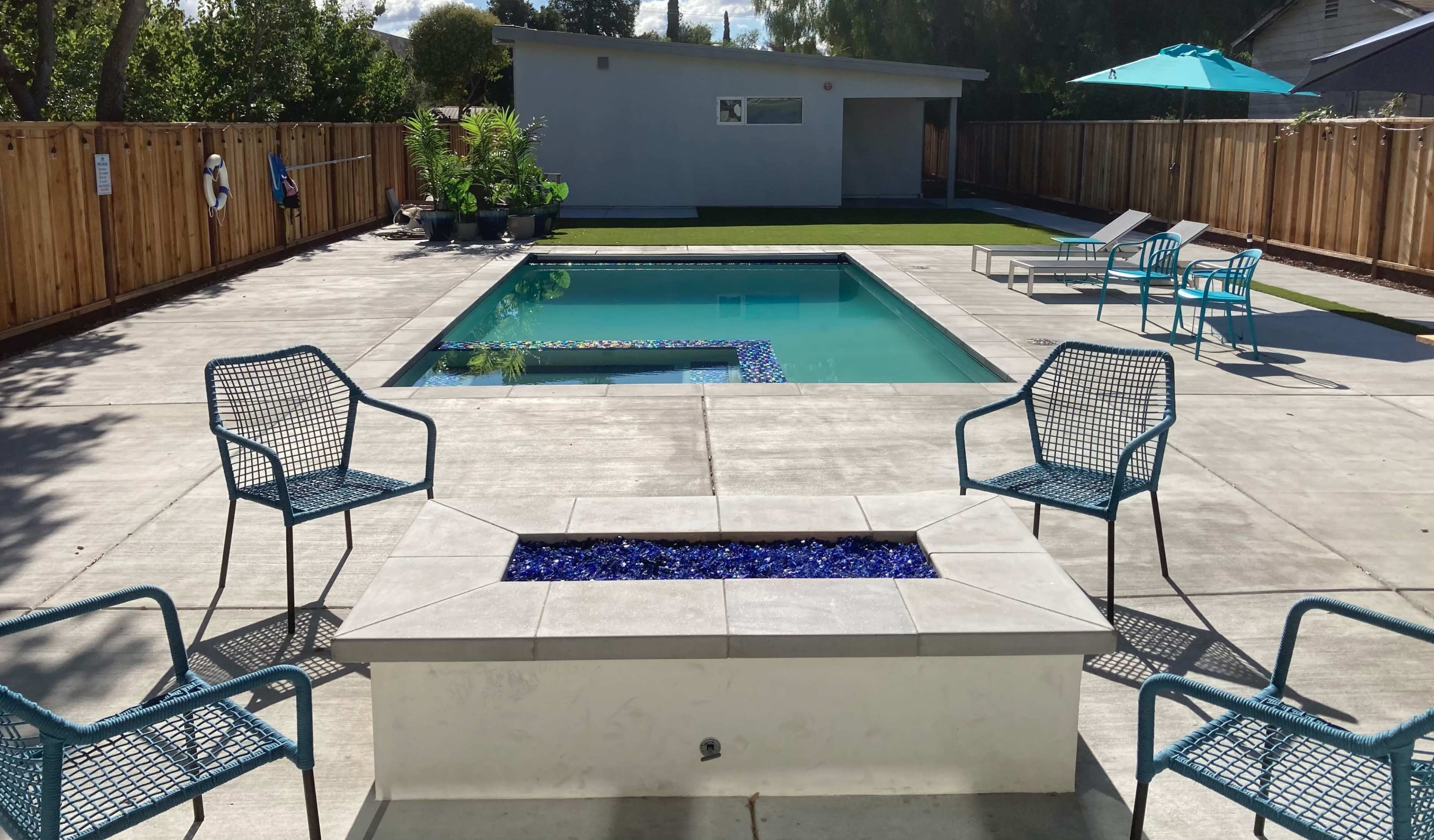 A swimming pool surrounded by concrete, with four blue chairs positioned around a fire pit and a small house in the background.