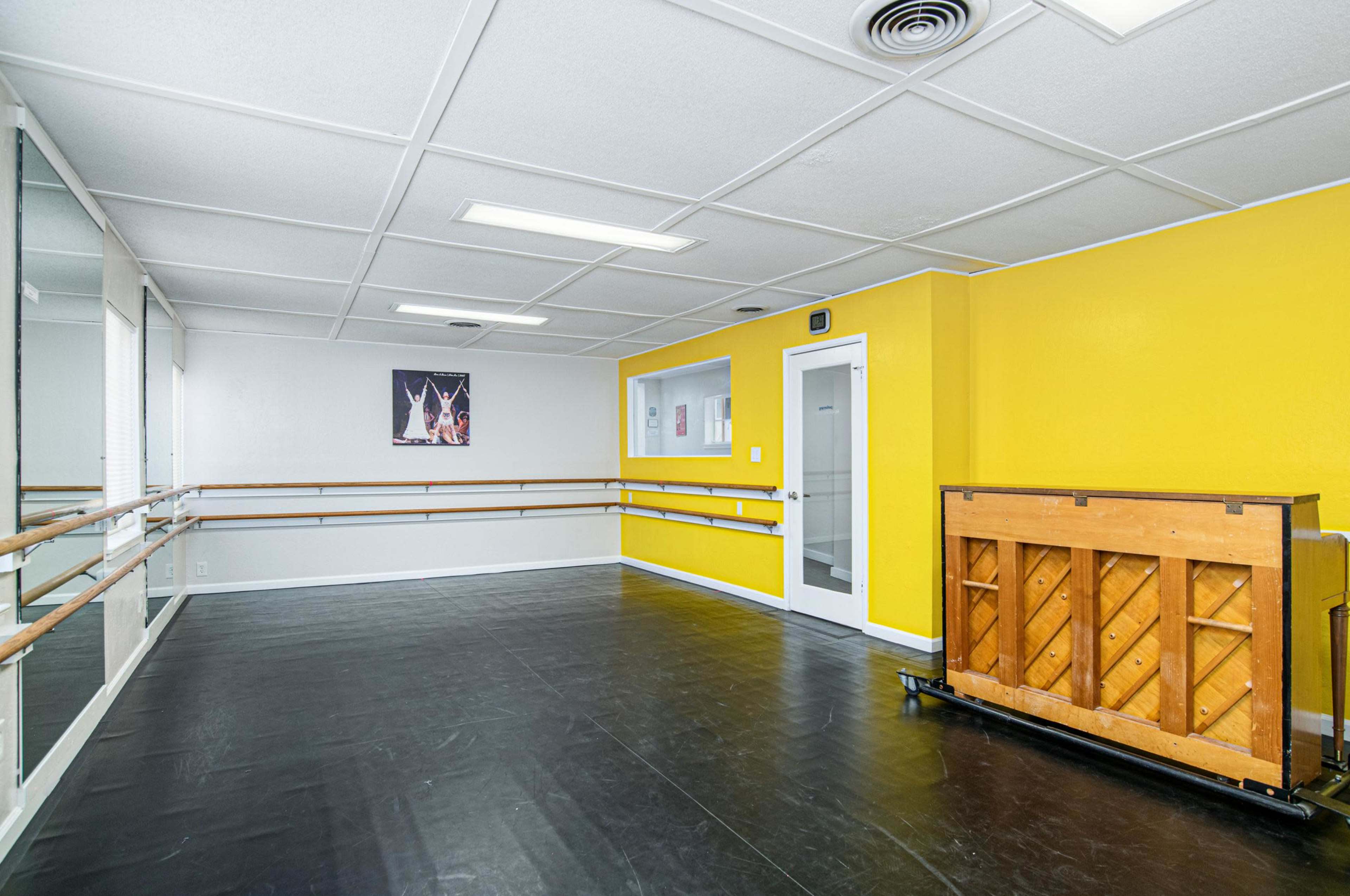 A spacious dance studio featuring a yellow wall, ballet barres along one side, and a wooden piano against another wall.