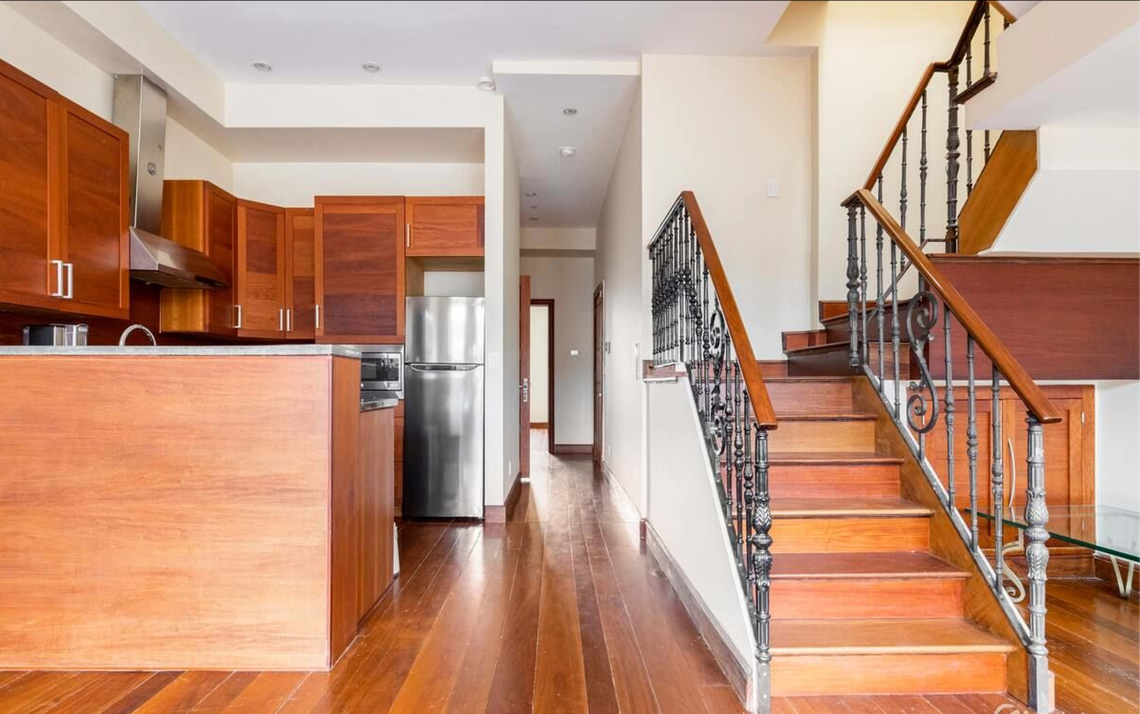 A modern kitchen area with wooden cabinets and a stainless steel refrigerator, leading to a wooden staircase.