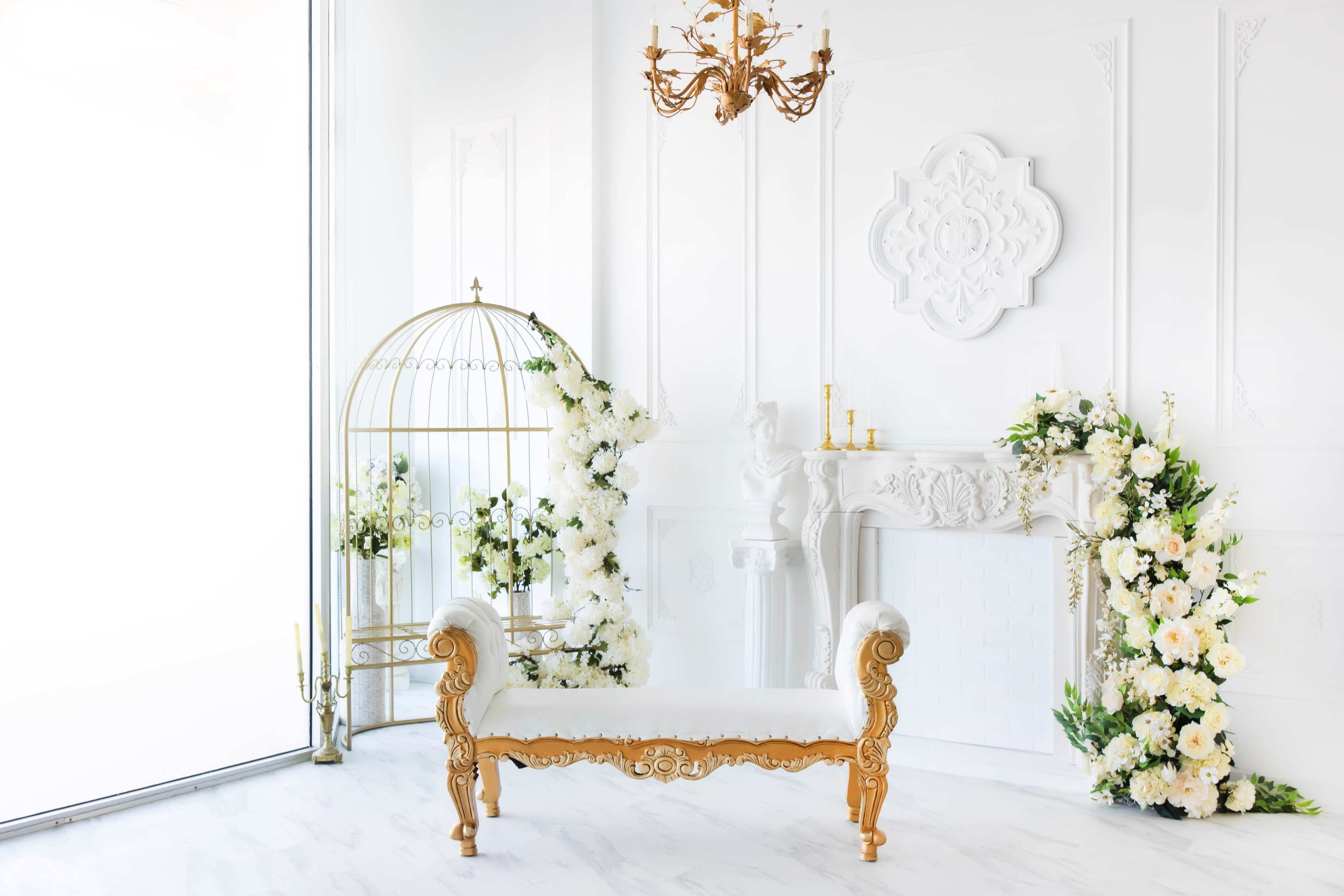 A bright, elegant room features a gold-accented bench, a birdcage, and a floral arrangement near a white mantelpiece.