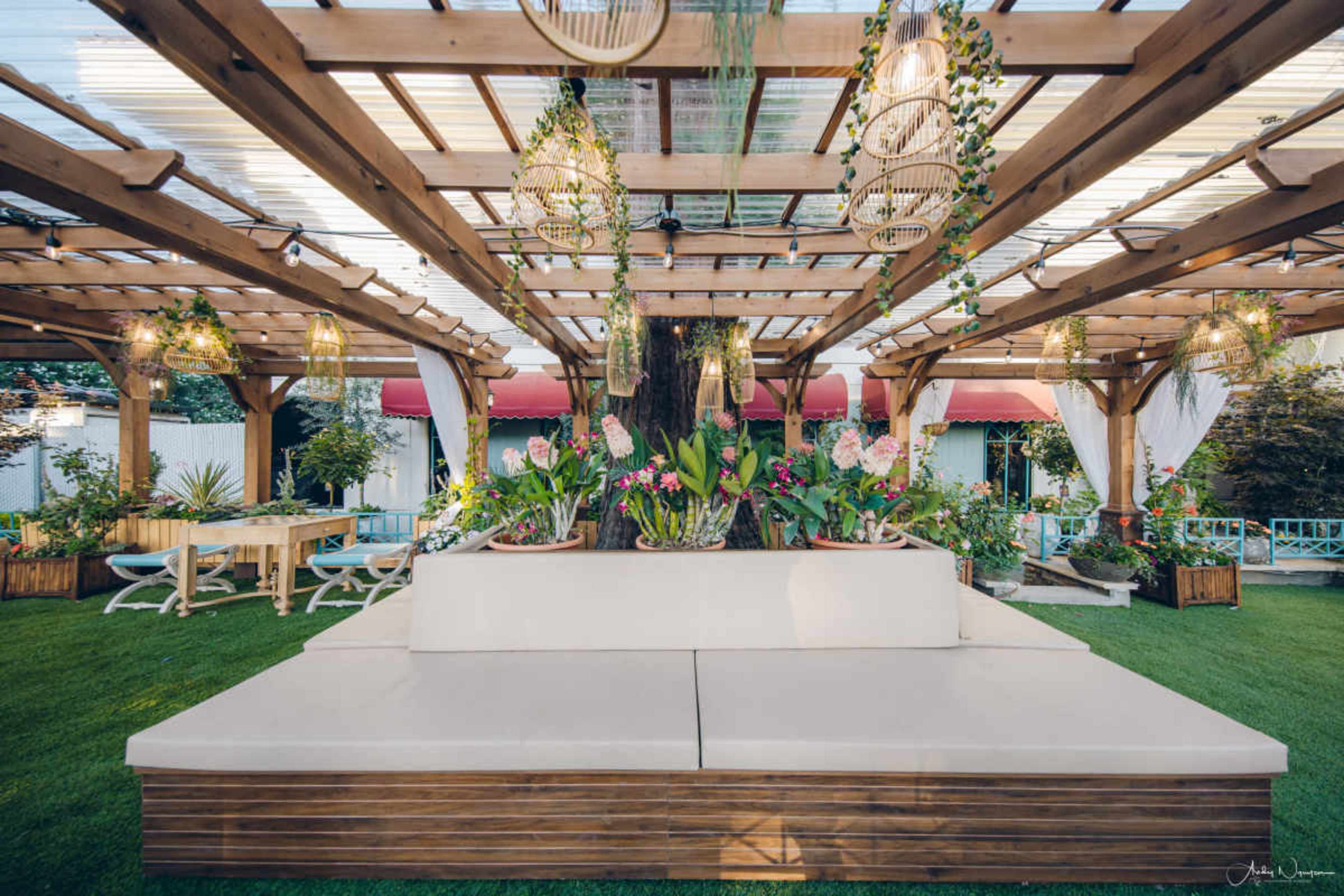 The image shows a landscaped outdoor space featuring a wooden pergola, a large, cushioned bench, and various potted plants and flowers.