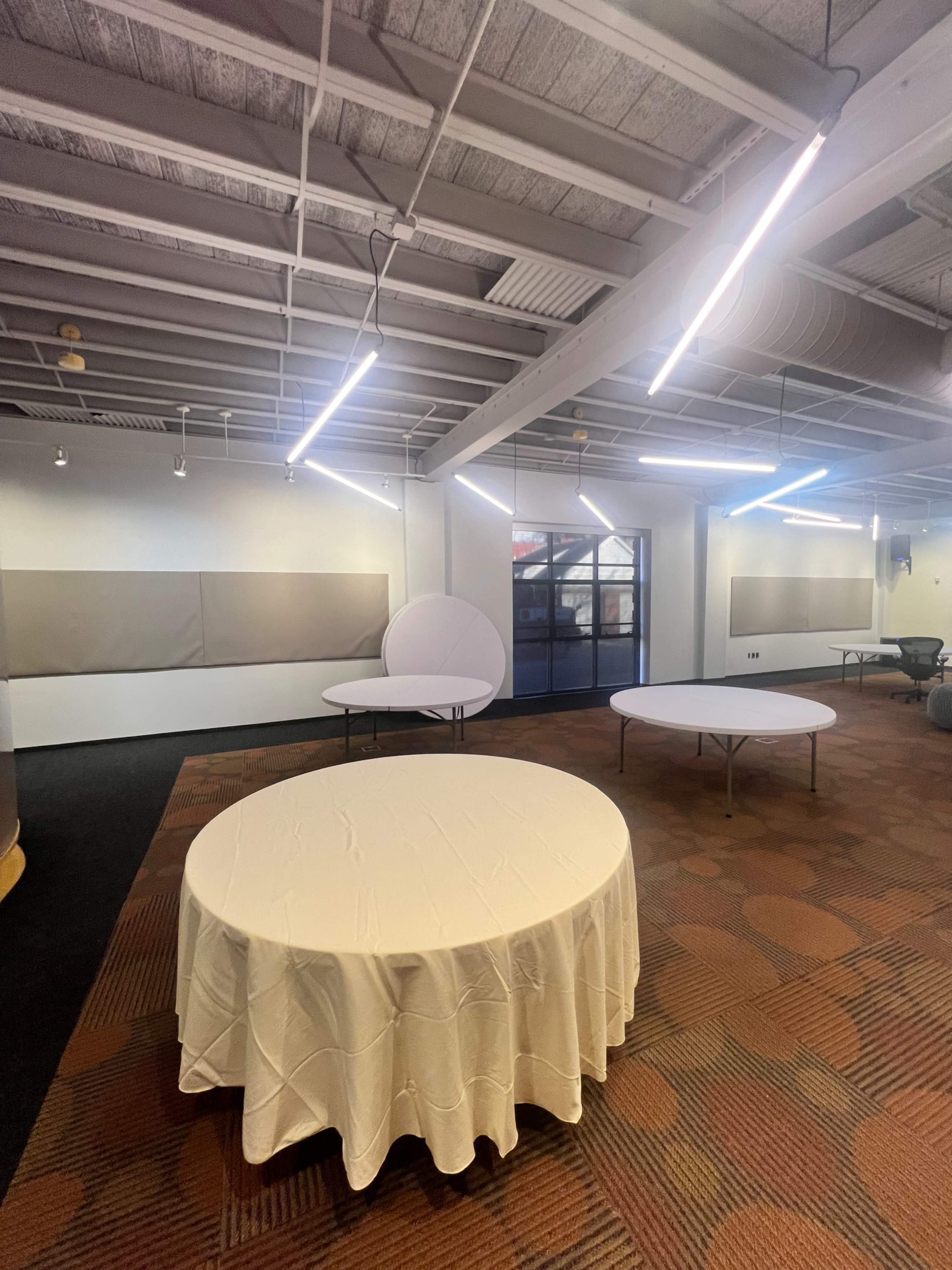 A spacious room with several round tables covered with white tablecloths, illuminated by overhead lights.