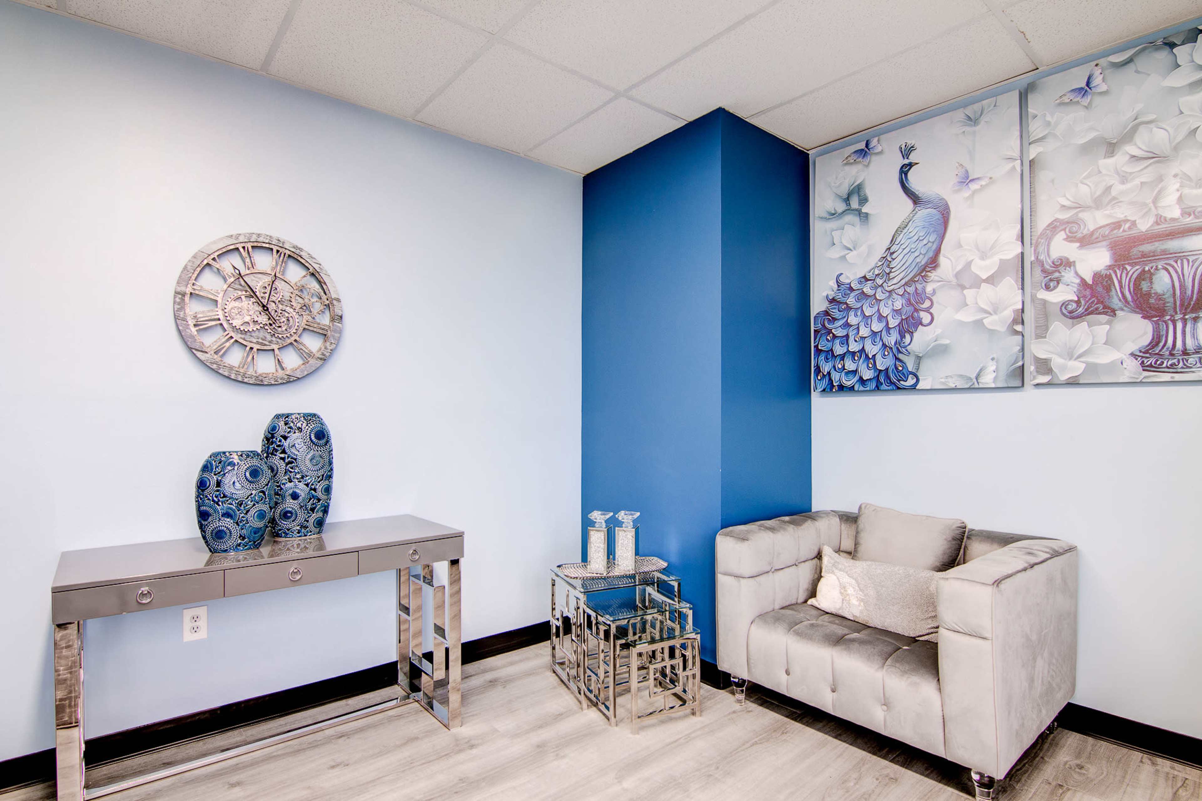The image shows a modern waiting area with a blue accent wall, a gray tufted sofa, a mirrored side table, and decorative wall art featuring a peacock.