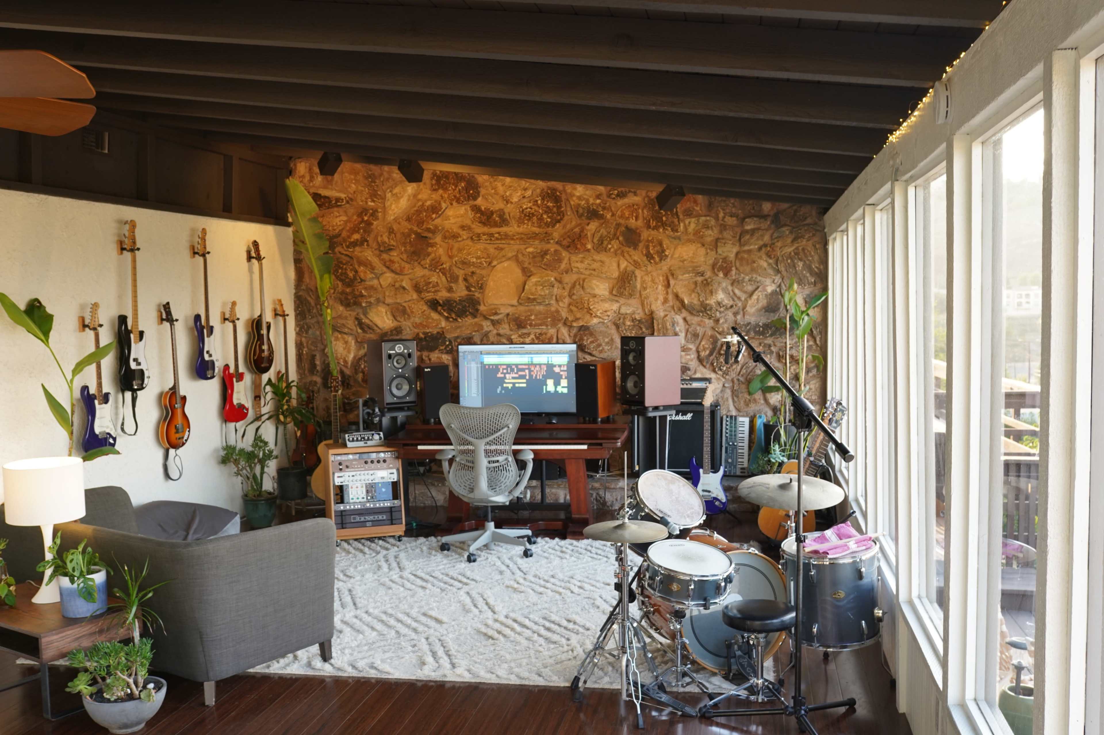 A music studio setup features a variety of instruments on the walls, a computer workstation, and a drum set in a well-lit room with large windows.