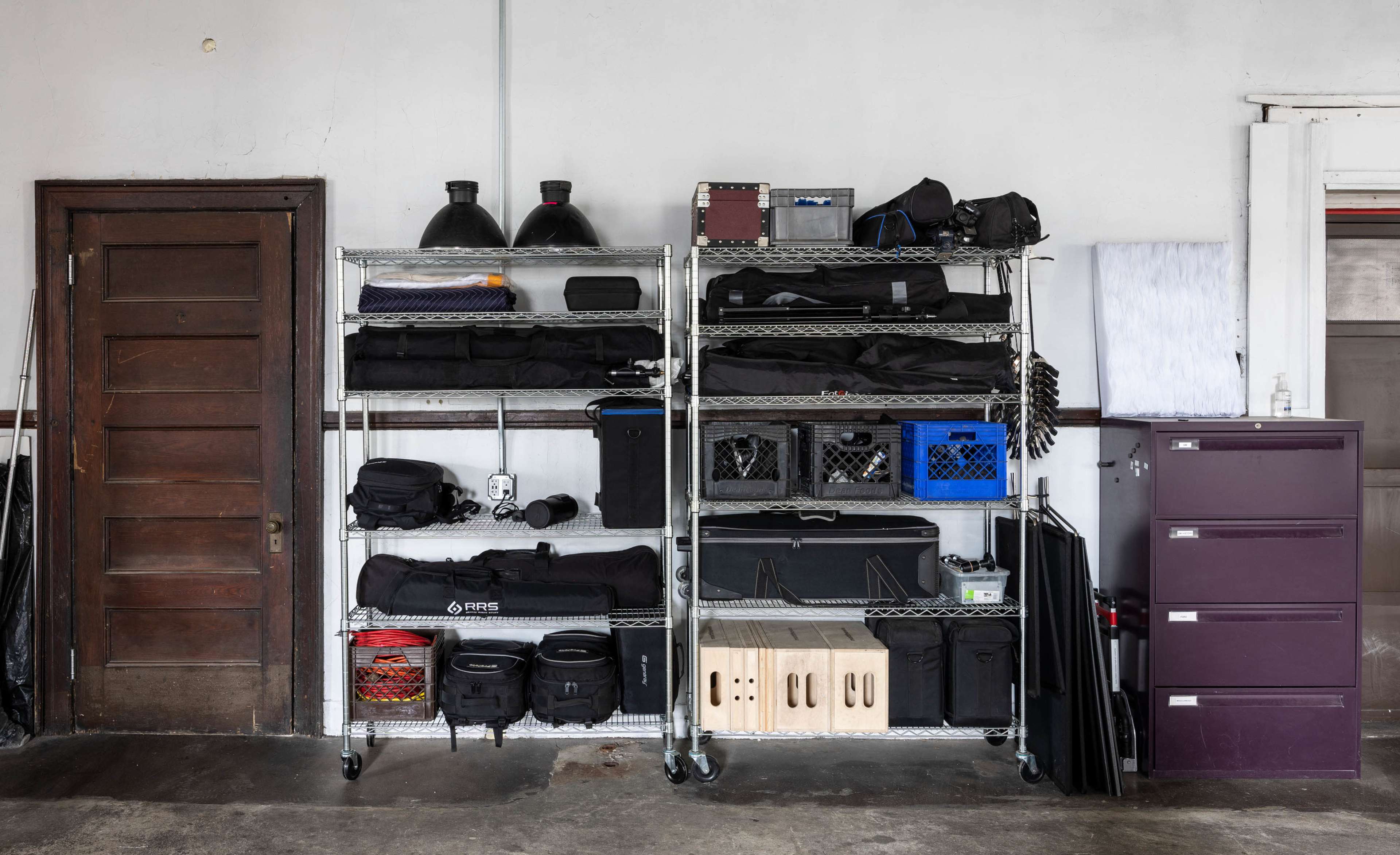 A metal shelving unit holds various storage bins, bags, and equipment in a garage-like space next to a brown door and a purple filing cabinet.