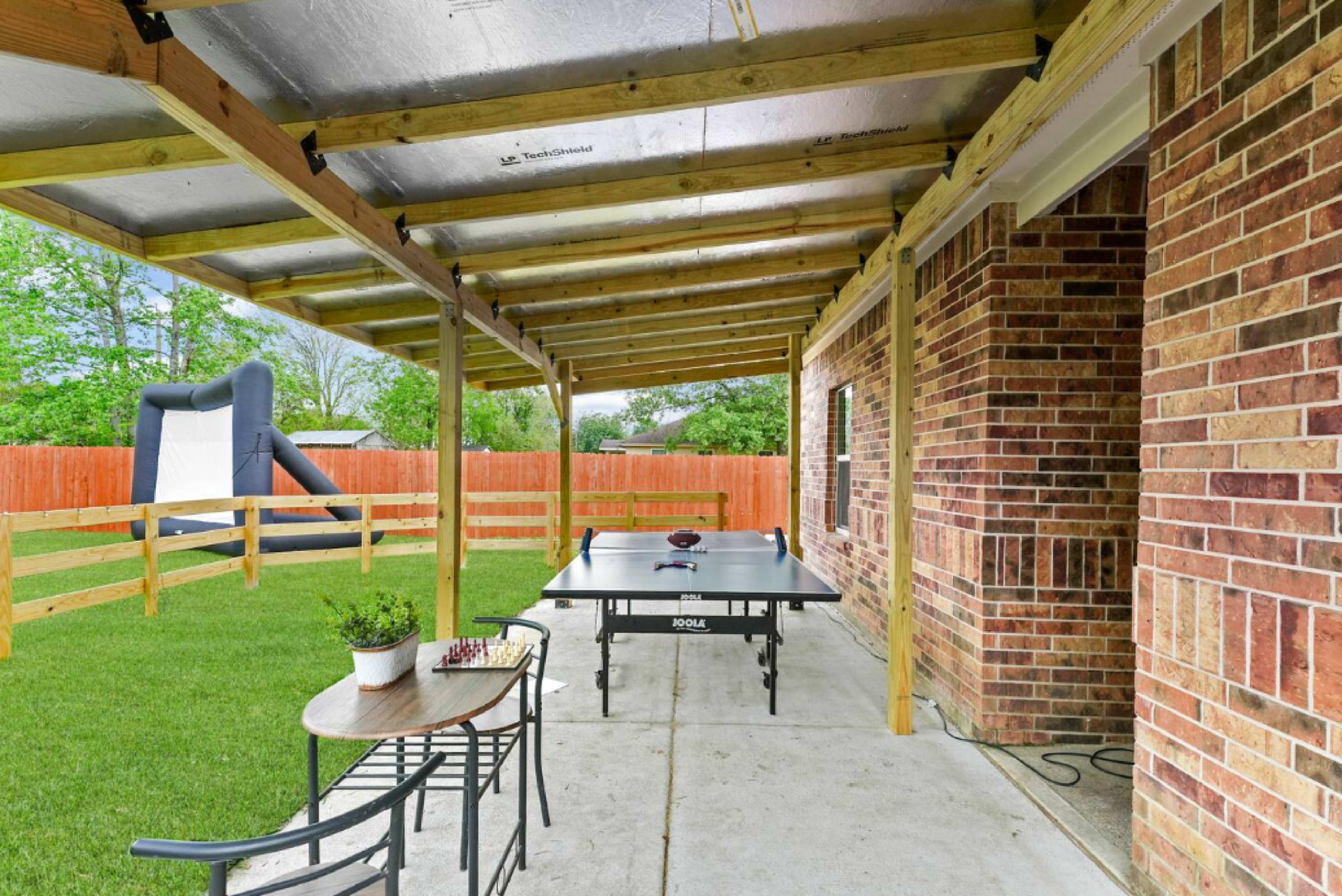 A covered patio features a ping pong table, a small table with a potted plant, and a fenced backyard with an inflatable bounce house.