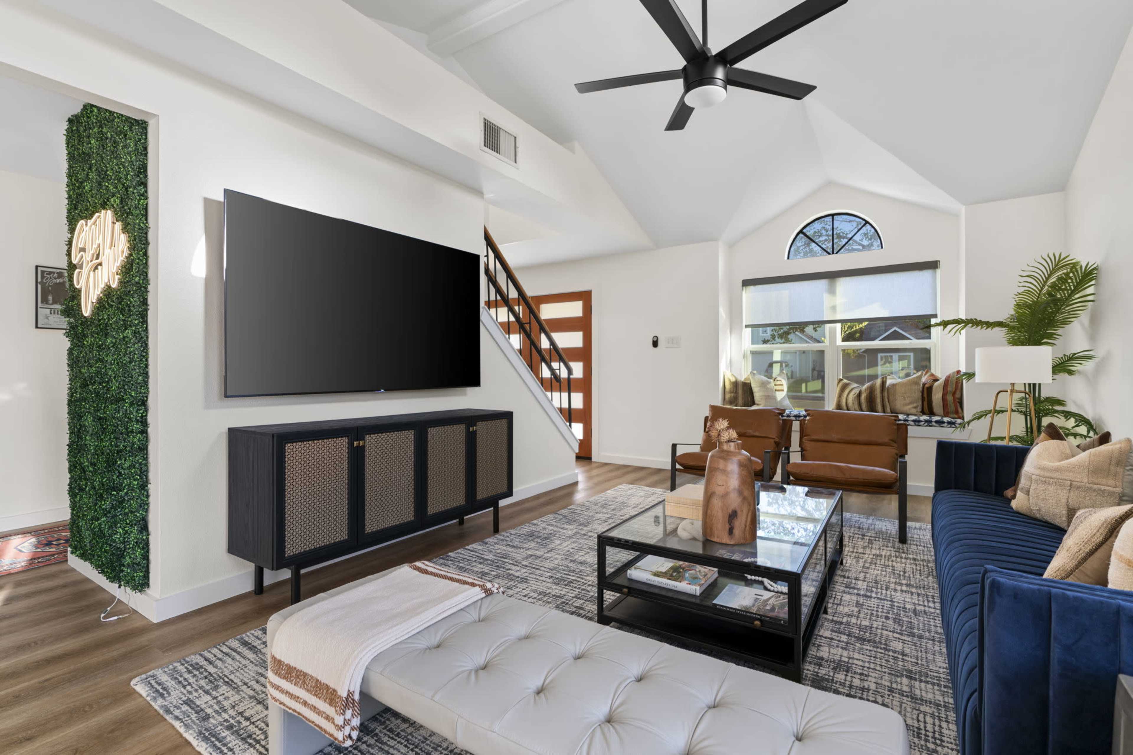 The living room features a large television mounted on a white wall, with a dark wooden cabinet underneath, a blue sectional sofa, brown armchairs, and a coffee table on a patterned rug.