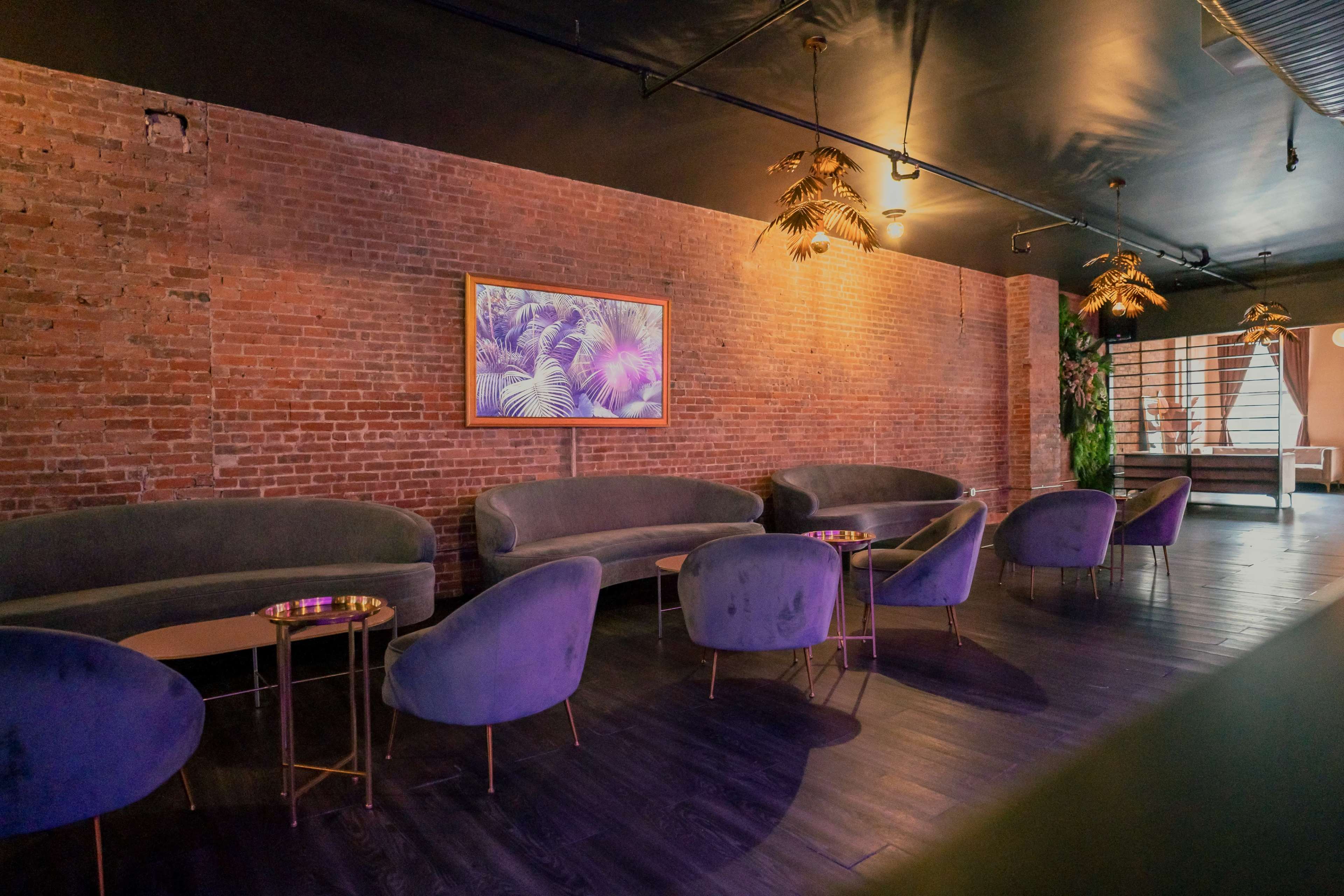The scene features a stylish lounge with purple upholstered seating, a brick wall, and decorative plants, complemented by framed artwork and low tables.