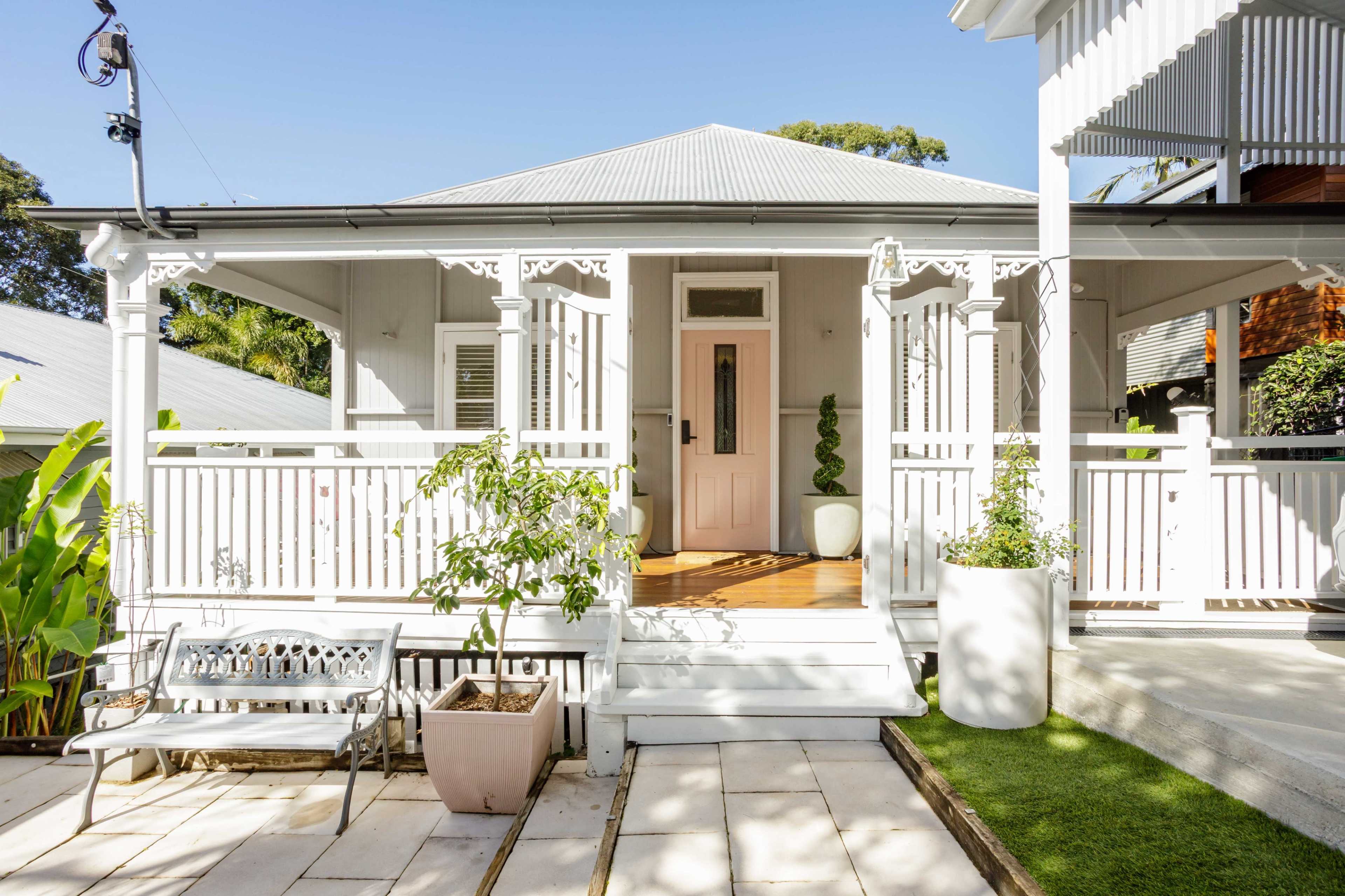 The image shows a quaint, white-painted house with a front porch adorned with decorative railing and potted plants.