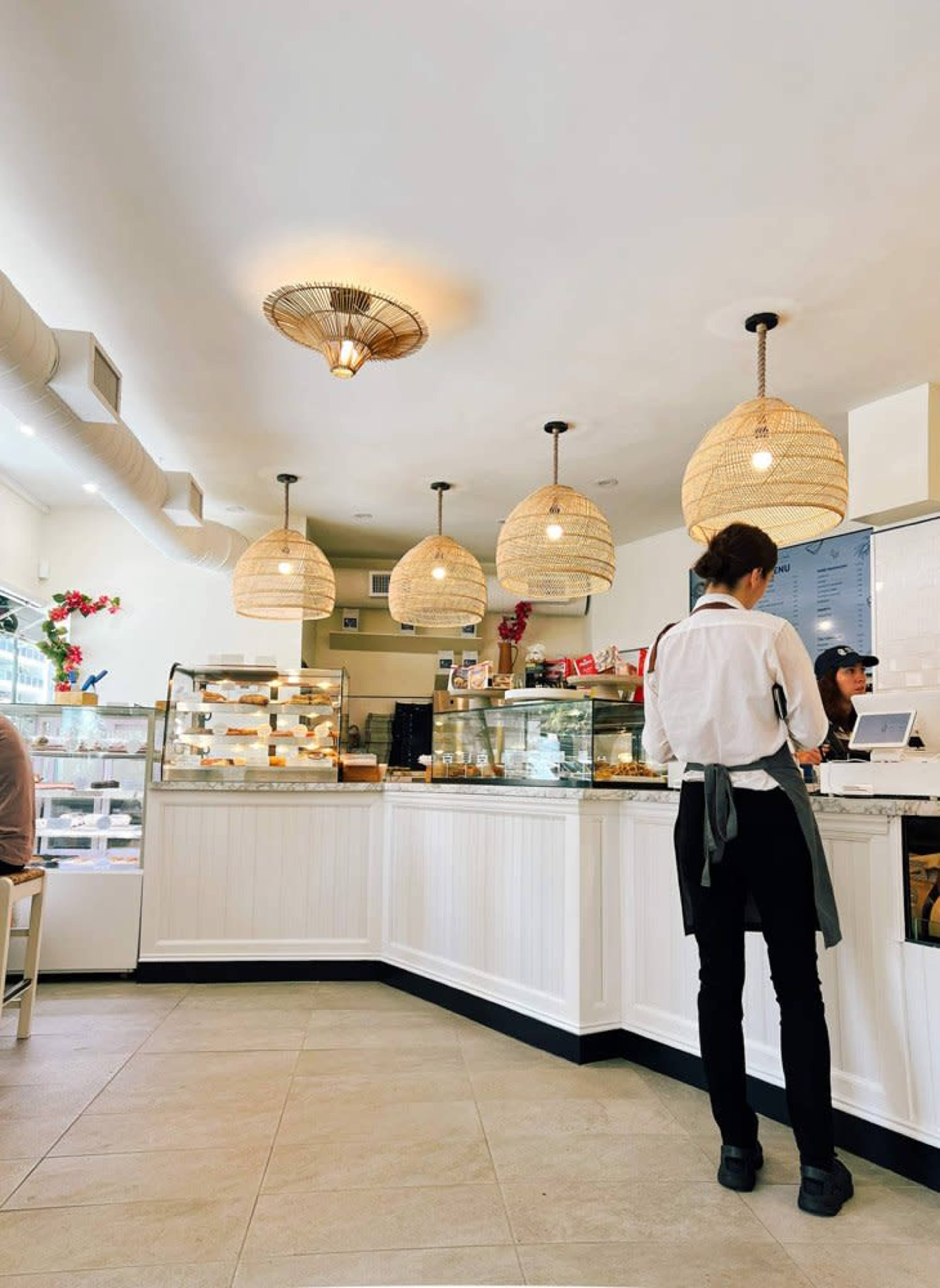 A café interior features a countertop display of desserts, a barista in a white shirt and apron, and hanging wicker light fixtures.