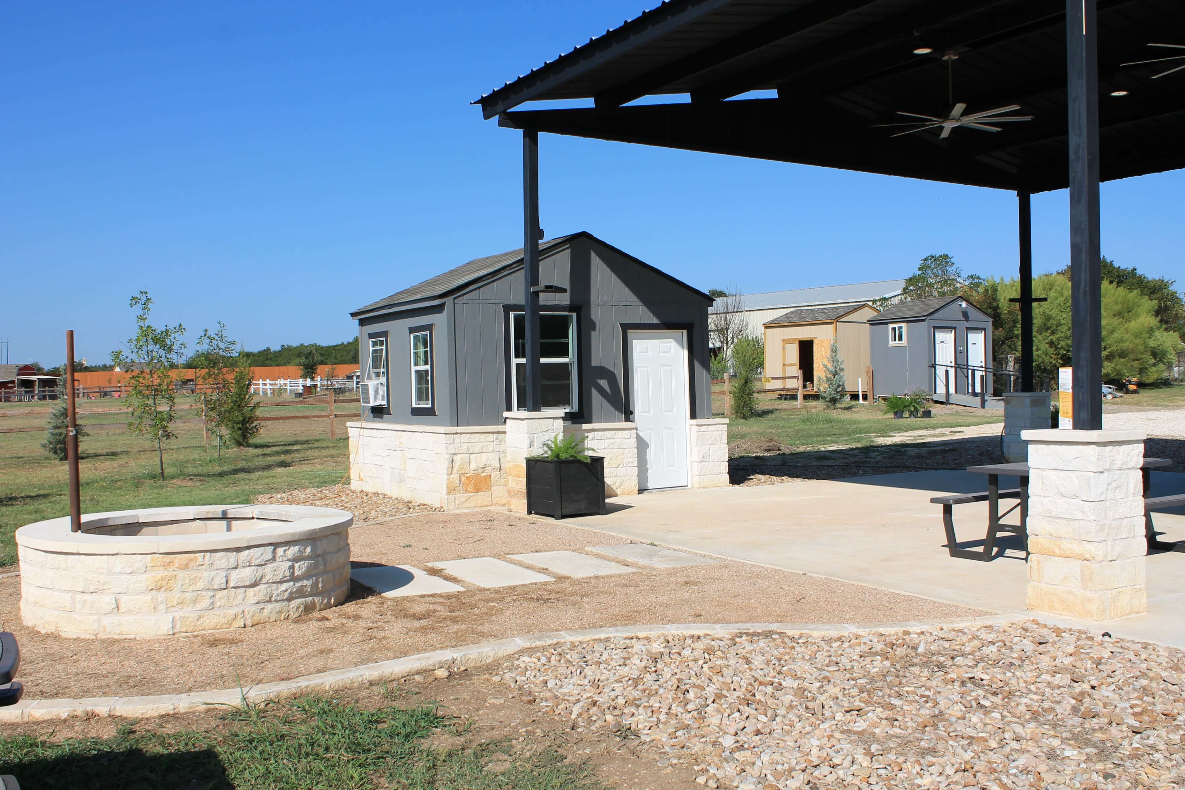 The image shows a small, modern shed beside a stone fire pit and a covered patio area in an open landscape with grass and distant structures.