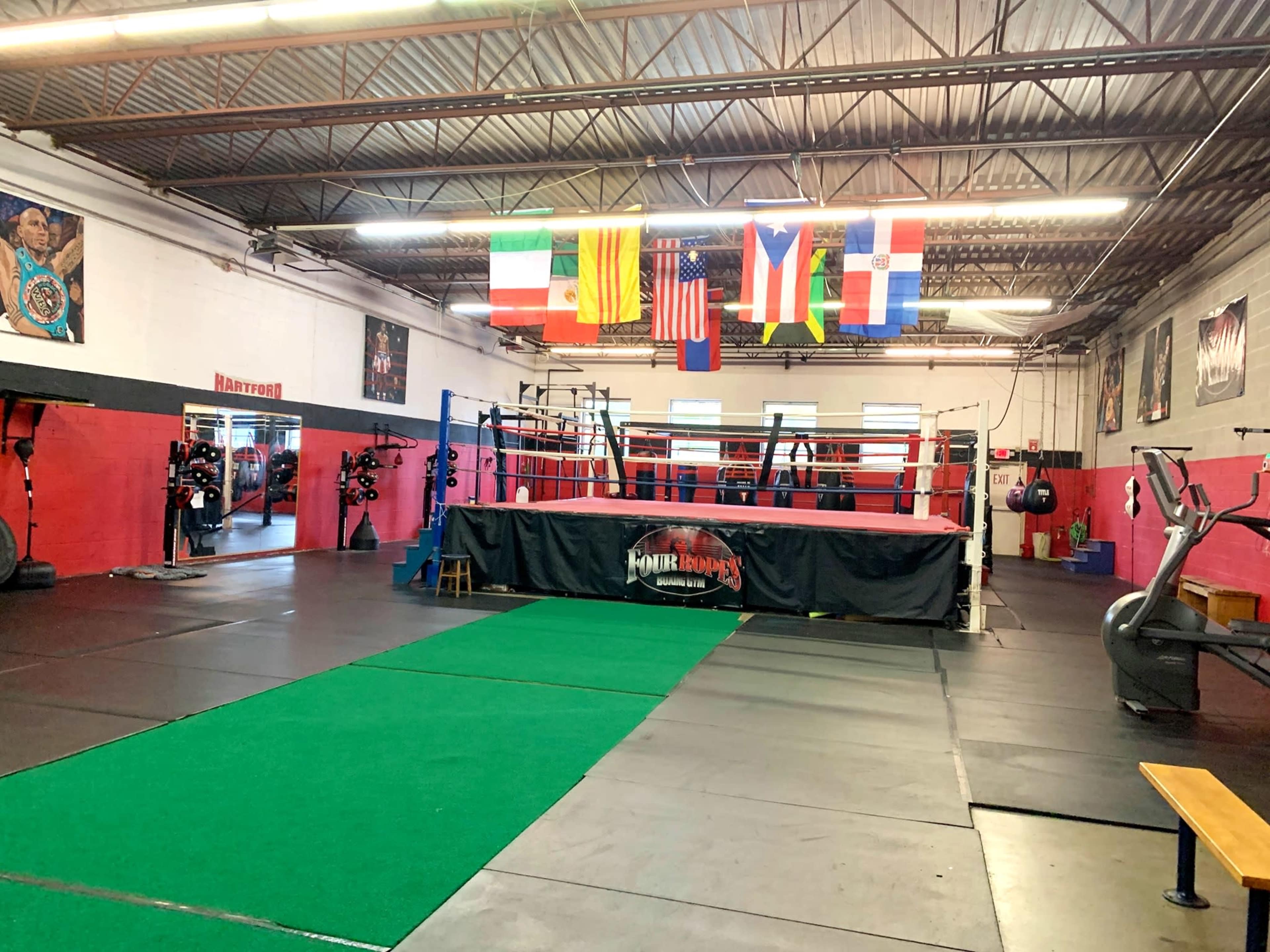 The image shows an indoor boxing gym with various flags hung from the ceiling, a boxing ring in the center, and exercise equipment on the sides.