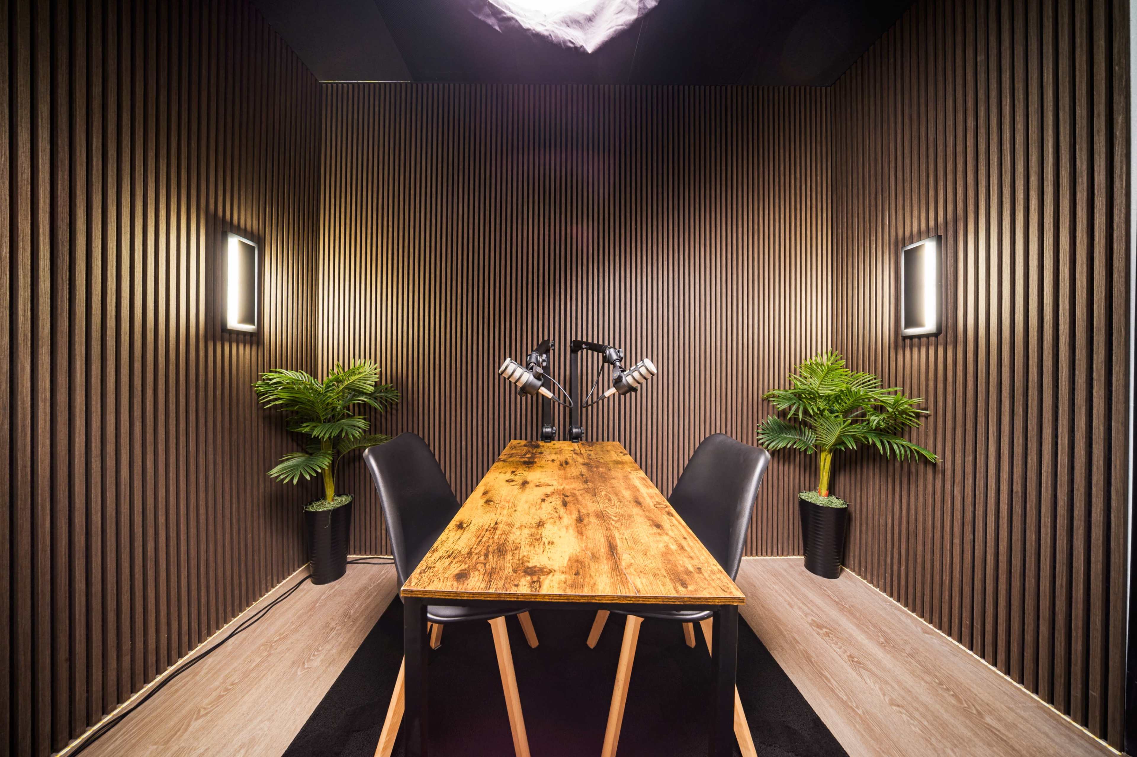 The image shows a modern podcasting studio with a wooden table, black chairs, and potted plants, surrounded by wooden paneling and soft lighting.