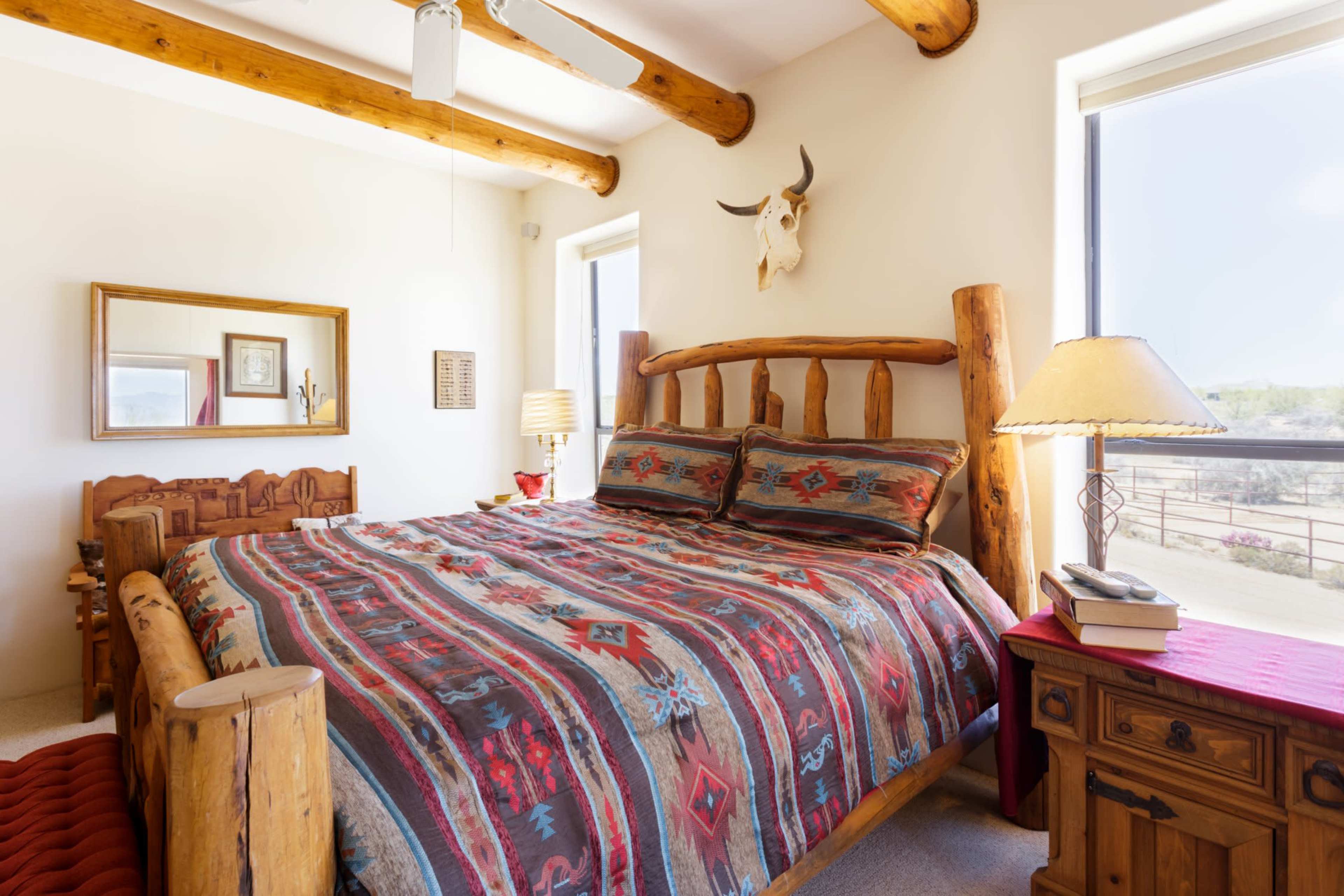 A rustic bedroom with a wooden bed frame, patterned bedding, and large windows allowing natural light to enter the space.