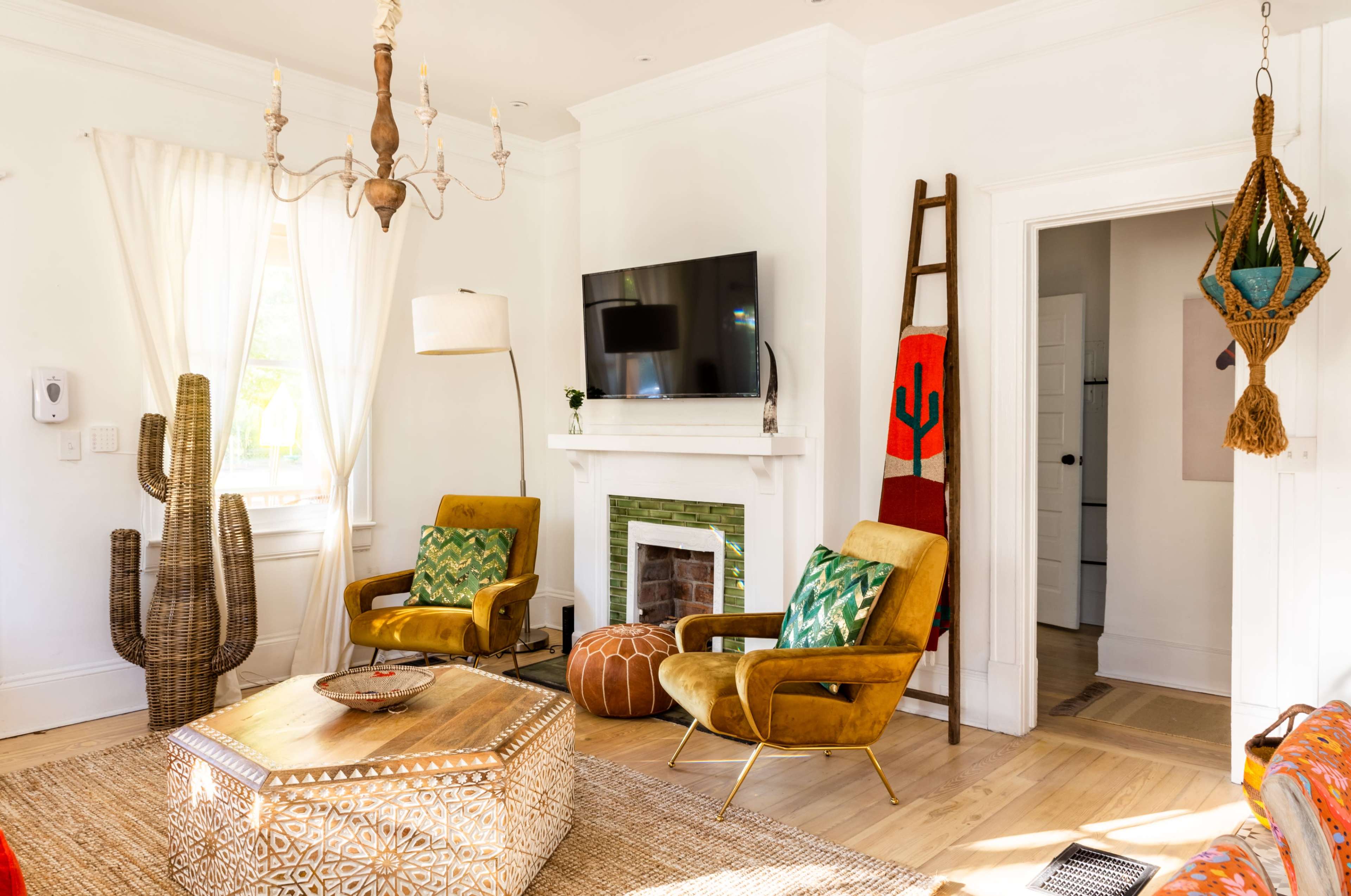 The living room features two yellow armchairs facing a coffee table, a television mounted above a white fireplace, and decorative items including a large cactus and a ladder.