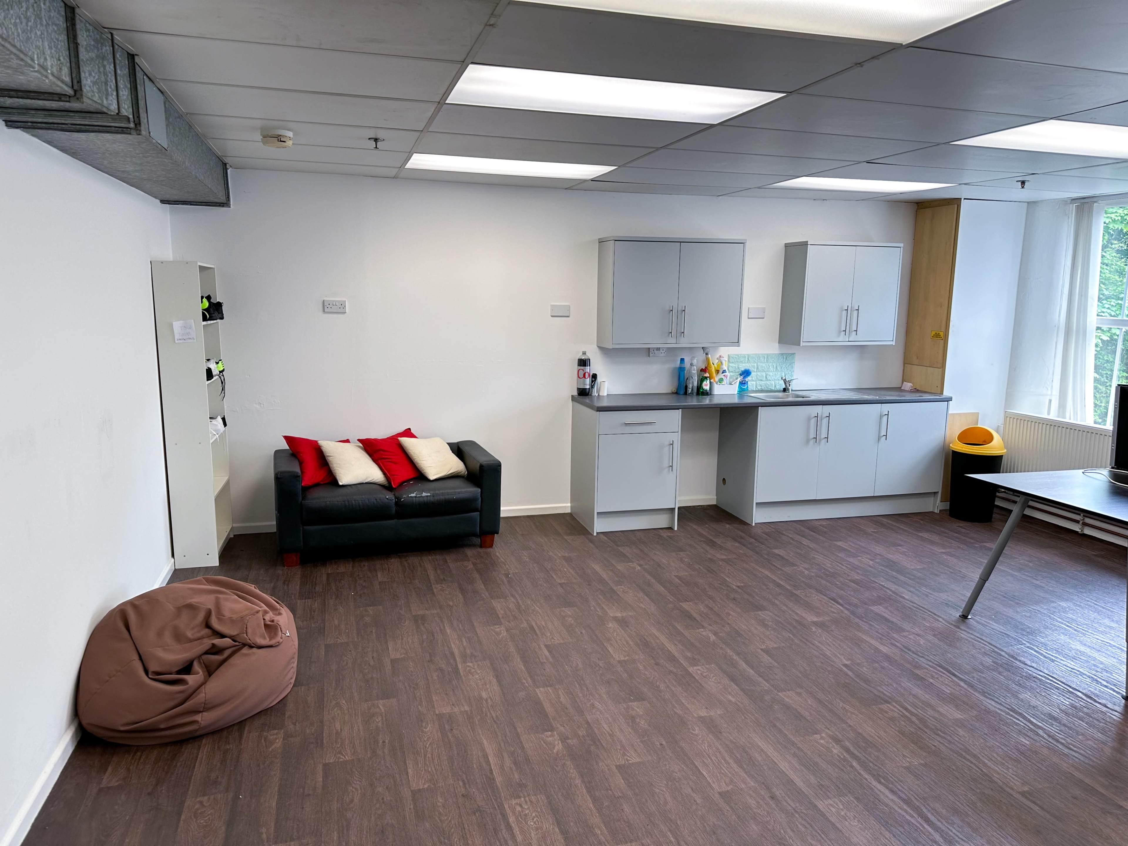 An office space features a couch with red cushions, a kitchenette with cabinets, and a beanbag chair on the floor.