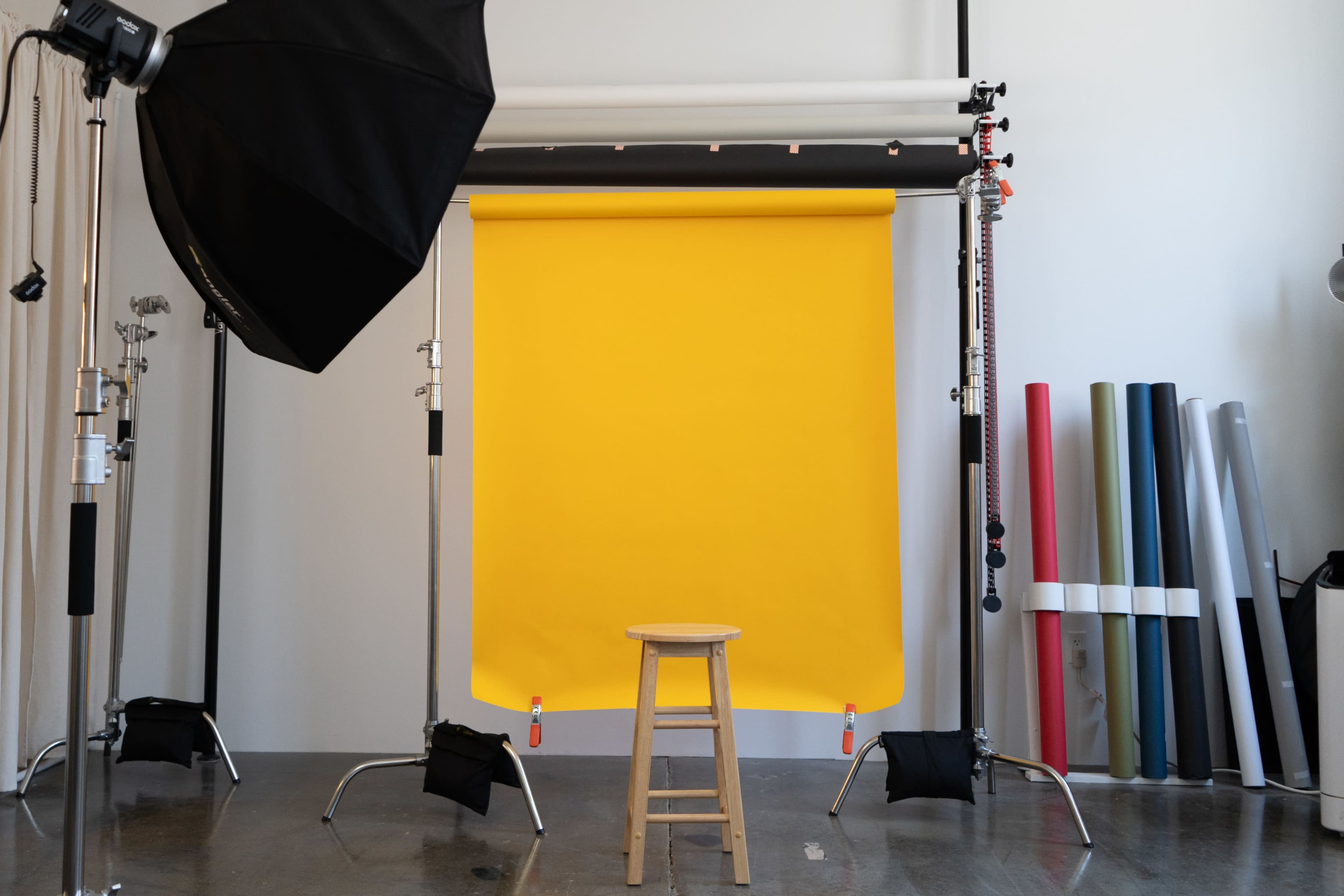 The image shows a photography studio setup featuring a yellow backdrop, a wooden stool in front of it, and various rolls of colored paper on the side.
