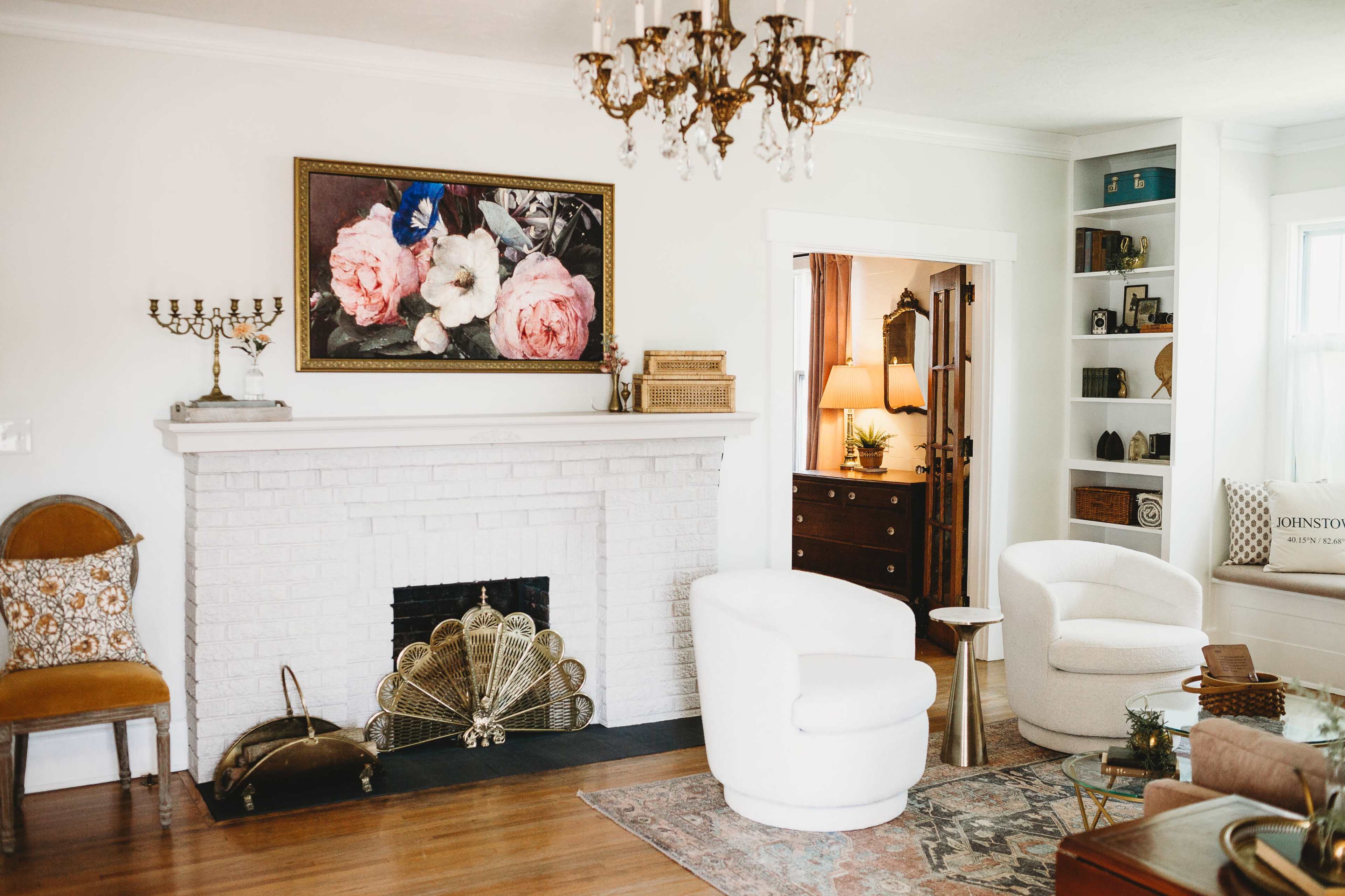 A stylish living room with a white fireplace, a large painting of flowers above it, and a mix of modern and vintage furniture, including a round white chair and a wooden side table.