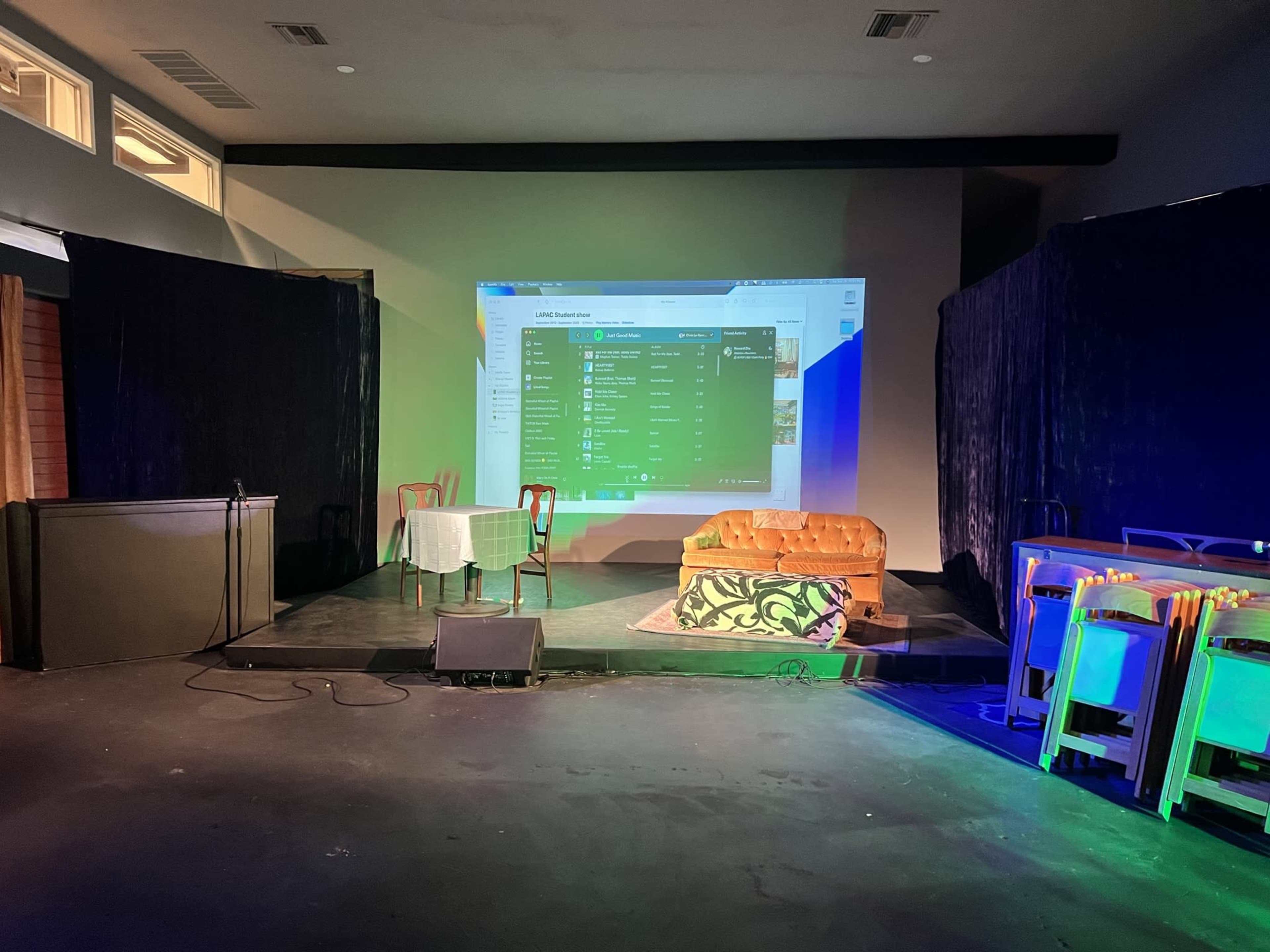 A stage setup with a large projection screen displaying a digital interface, a table with a chair, a sofa, and some furniture under soft lighting.