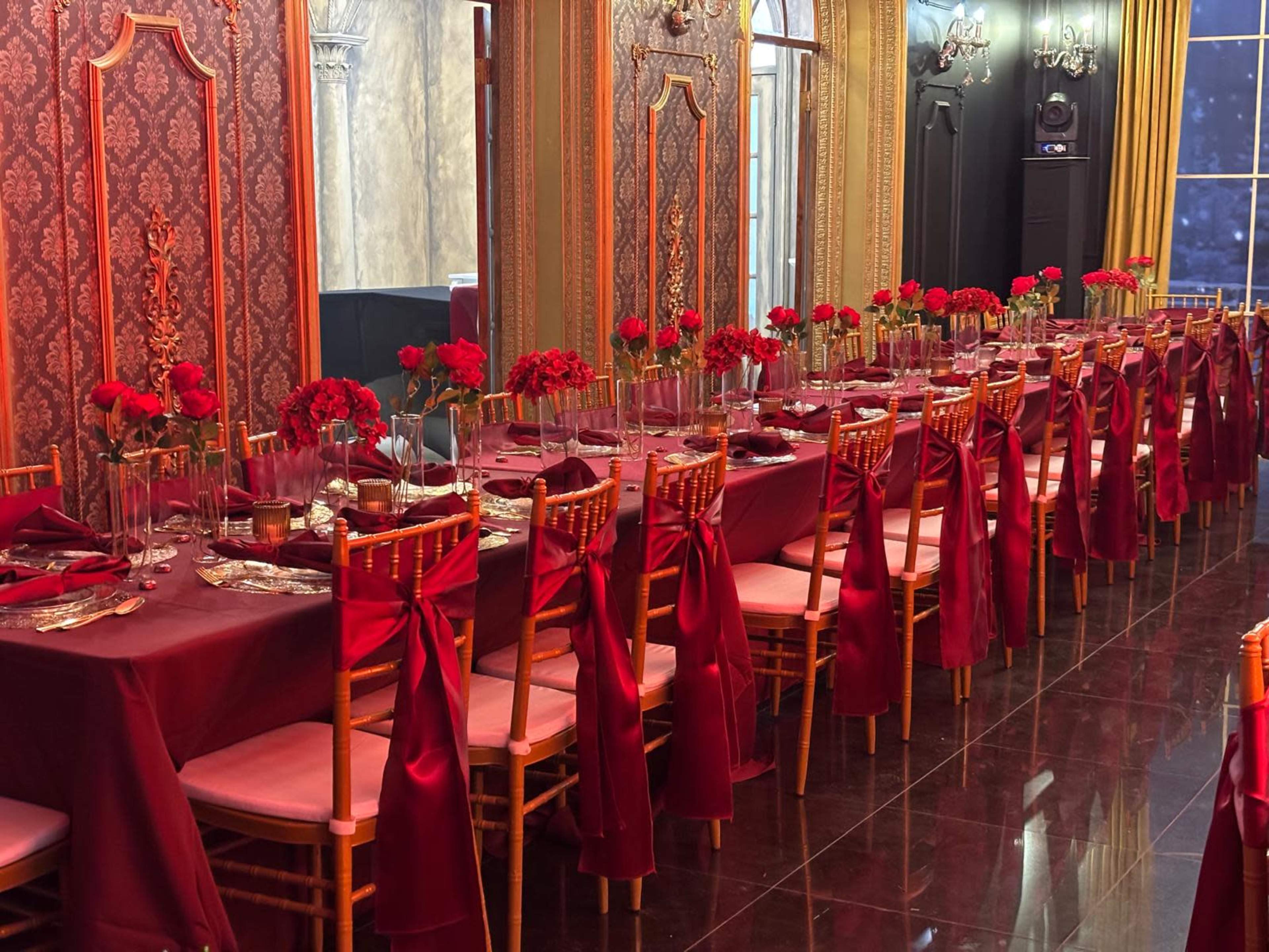 A long banquet table is elegantly set with red roses and gold tableware, surrounded by chiavari chairs adorned with burgundy sashes in a luxurious dining hall.
